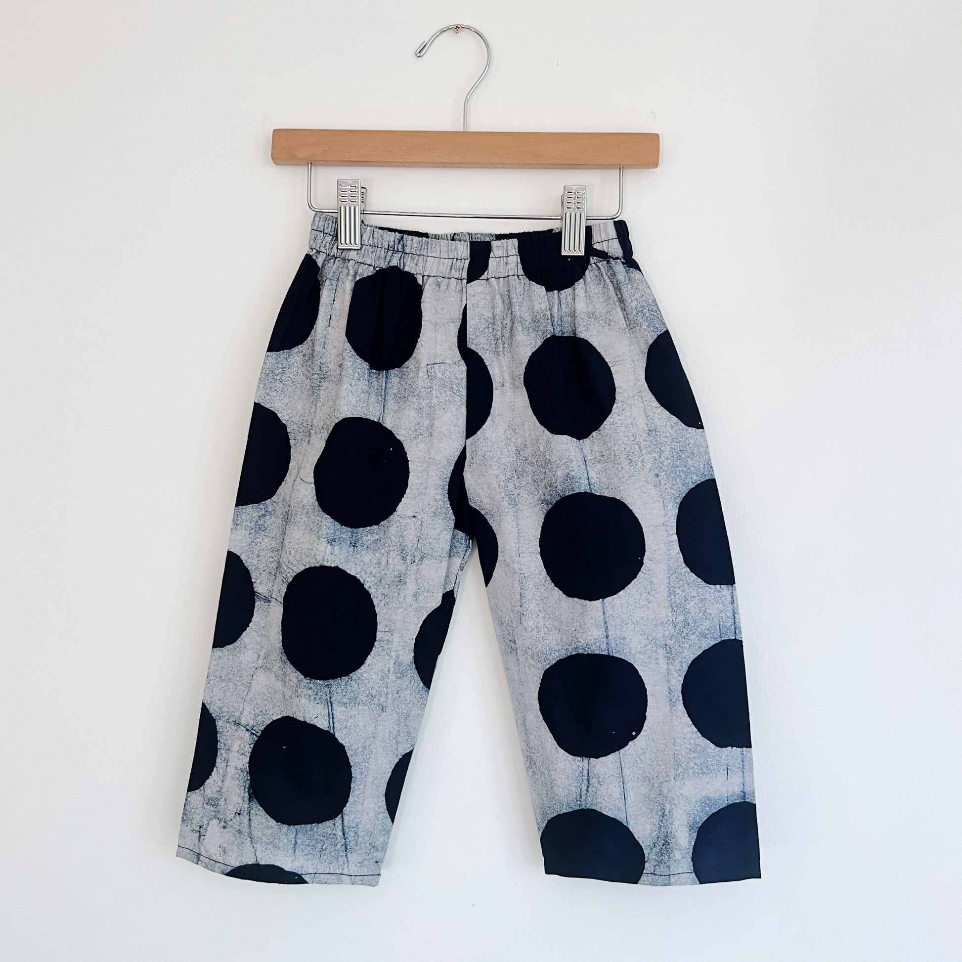 Kids lazy pants in black dots Batiki cotton, hanging flat on a wooden clip hanger against a white wall, showing elasticated waistband, oversized hand-printed black polka dots on a grey textured ground, and relaxed fit