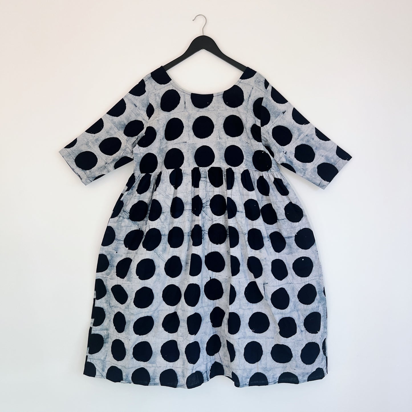 Full front view of the Sula Dress in Black Indigo Dots, hanging on a black hanger against a white wall, showing a round neckline, elbow-length sleeves, gathered waist seam, and oversized hand-printed black polka dots on grey-white 100% cotton Batiki fabric

