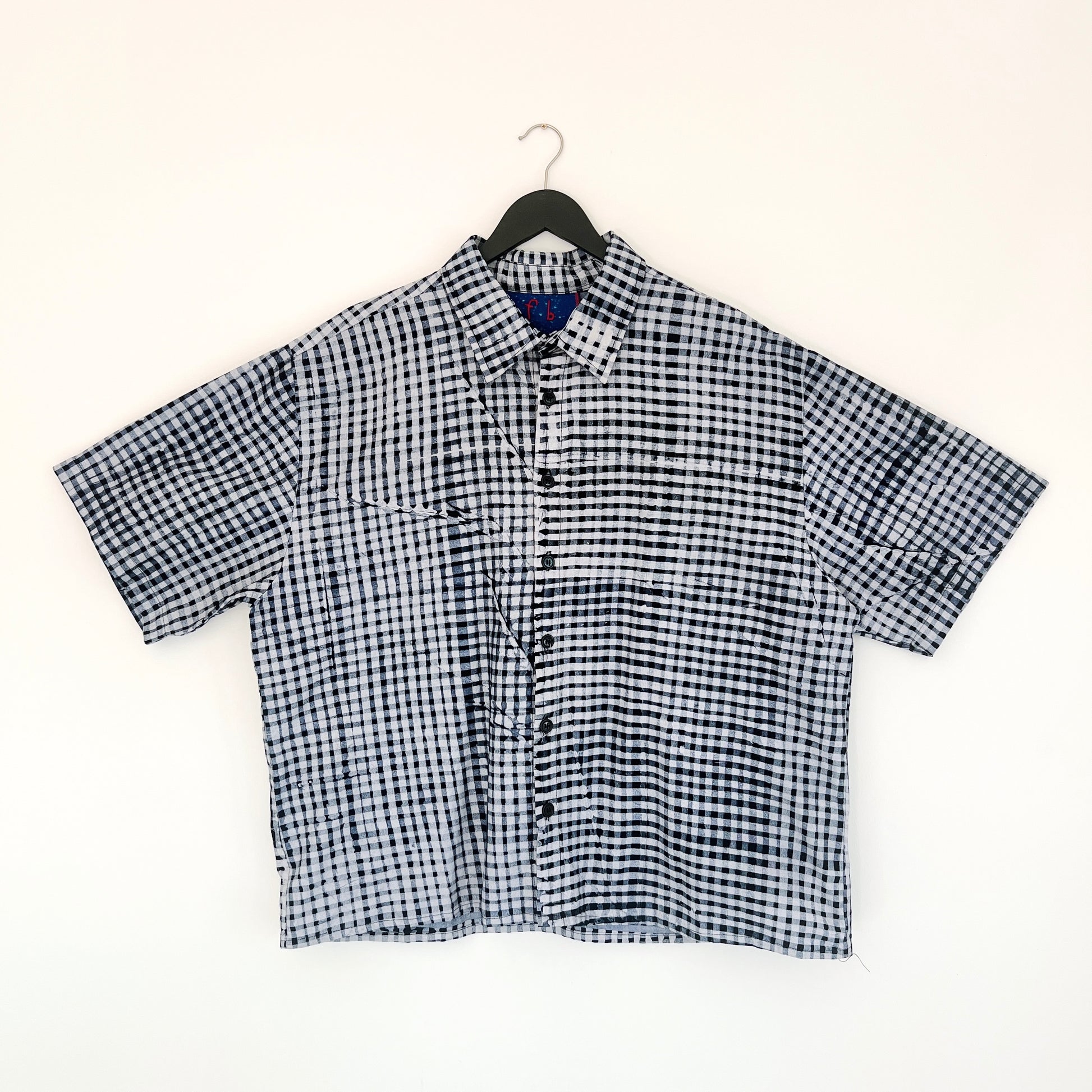 Full front view of the Sudi Shirt in Indigo Grid, hanging on a black hanger against a white wall, showing a point collar, short sleeves, button placket, two chest pockets, and hand-printed indigo and white gingham grid pattern on 100% cotton Batiki fabric

