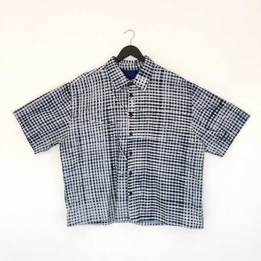 Full front view of the Sudi Shirt in Indigo Grid, hanging on a black hanger against a white wall, showing a point collar, short sleeves, button placket, two chest pockets, and hand-printed indigo and white gingham grid pattern on 100% cotton Batiki fabric

