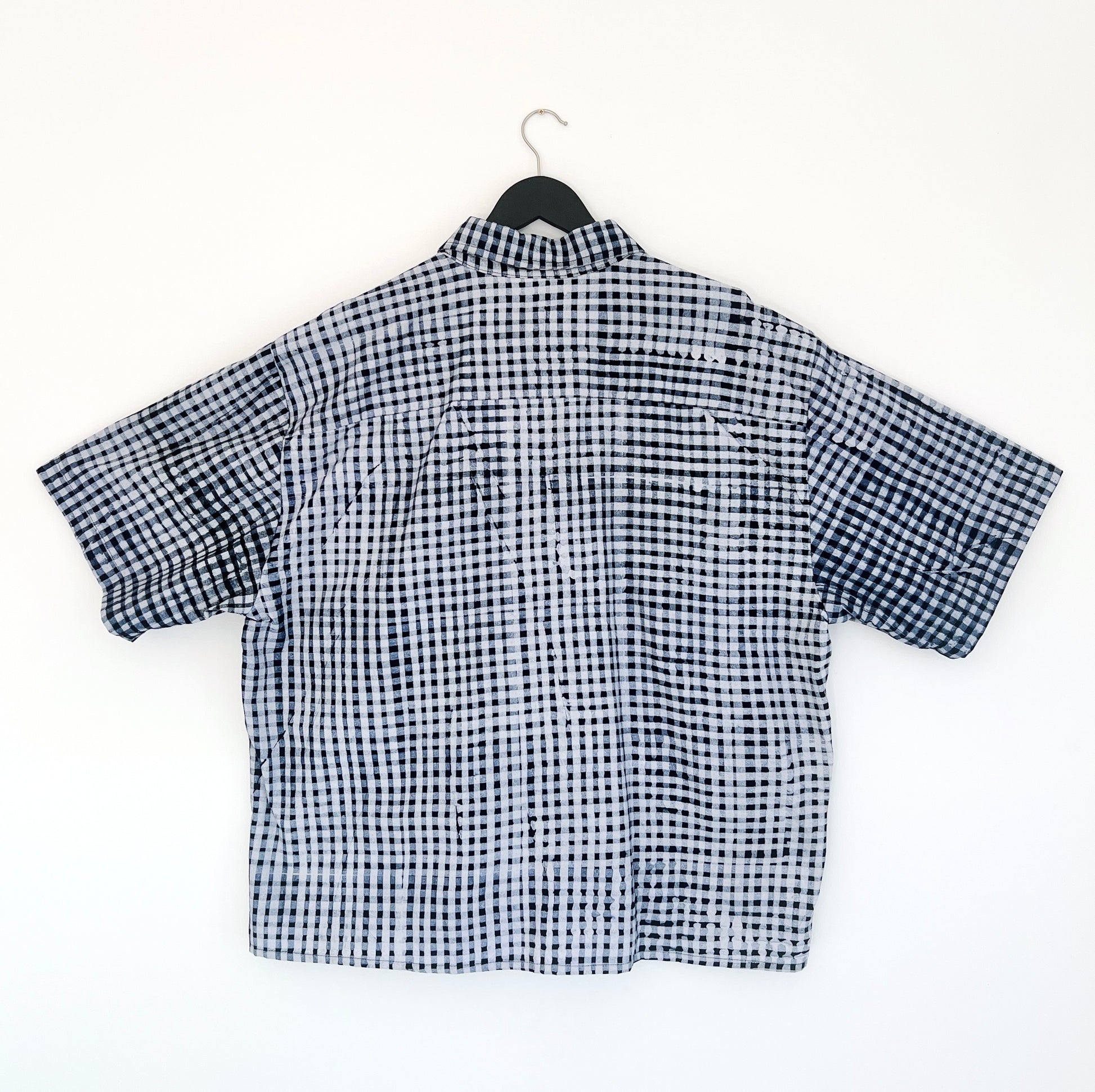 Back view of the Sudi Shirt in Indigo Grid, hanging on a black hanger against a white wall, showing the relaxed boxy silhouette, short sleeves, back yoke seam, and hand-printed indigo and white gingham grid pattern on 100% cotton Batiki fabric

