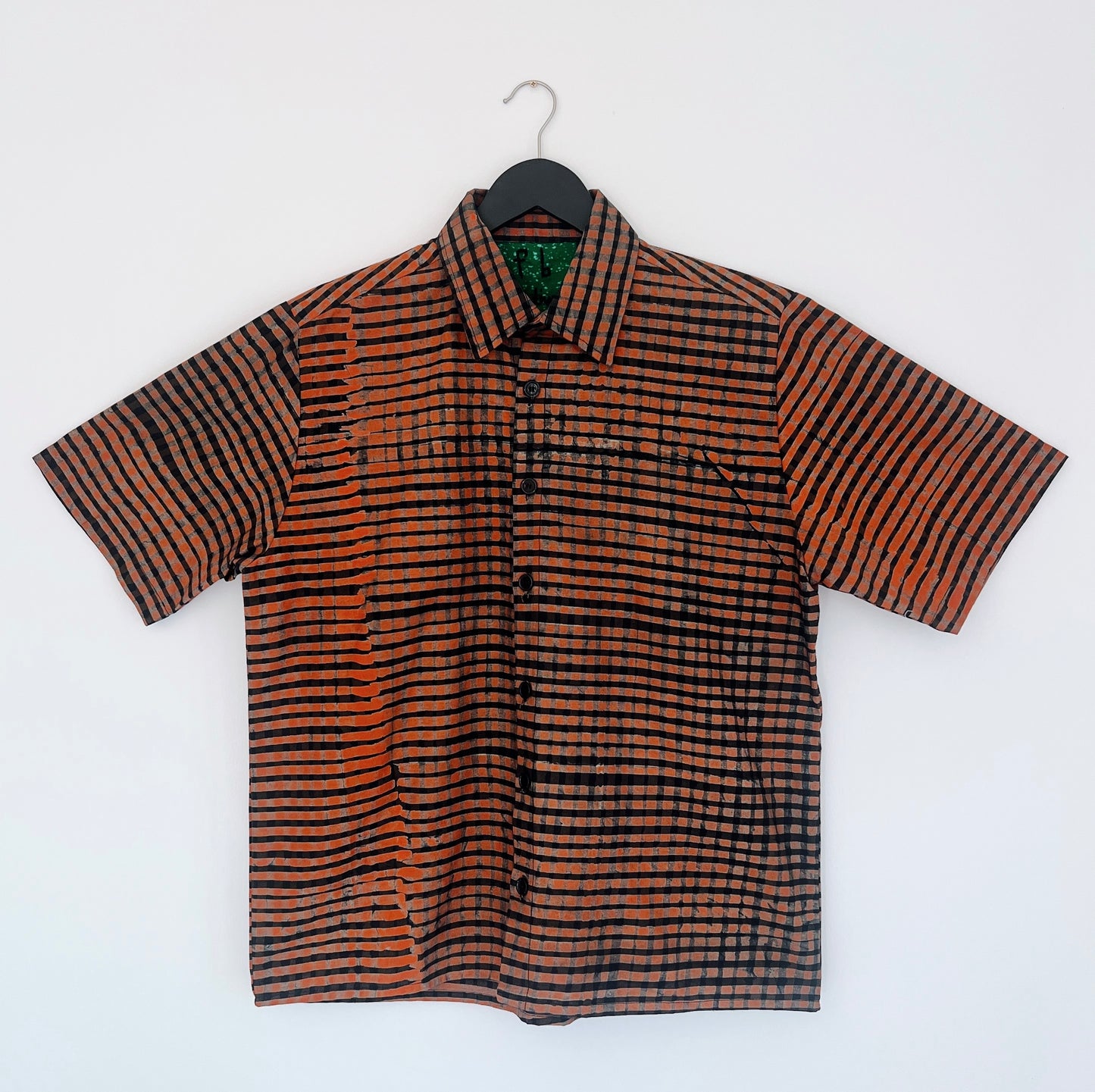 Full front view of the Sudi Shirt in Persimmon Grid, hanging on a black hanger against a white wall, showing a point collar, short sleeves, button placket, and hand-printed persimmon orange and black grid check pattern on 100% cotton Batiki fabric with green patterned collar lining

