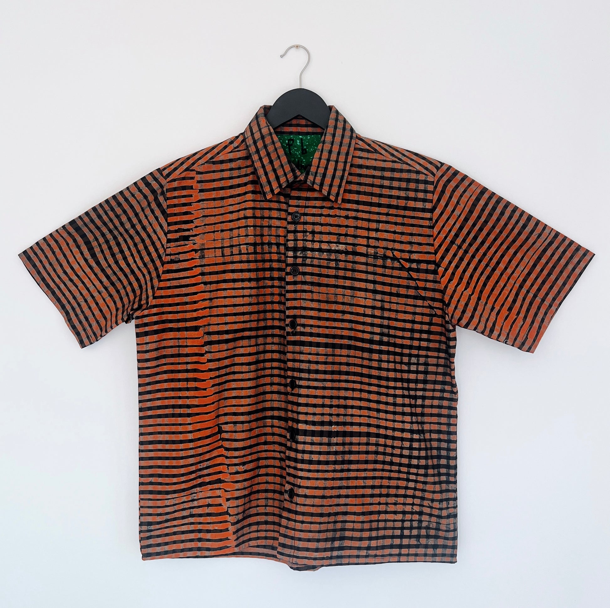 Full front view of the Sudi Shirt in Persimmon Grid, hanging on a black hanger against a white wall, showing a point collar, short sleeves, button placket, and hand-printed persimmon orange and black grid check pattern on 100% cotton Batiki fabric with green patterned collar lining

