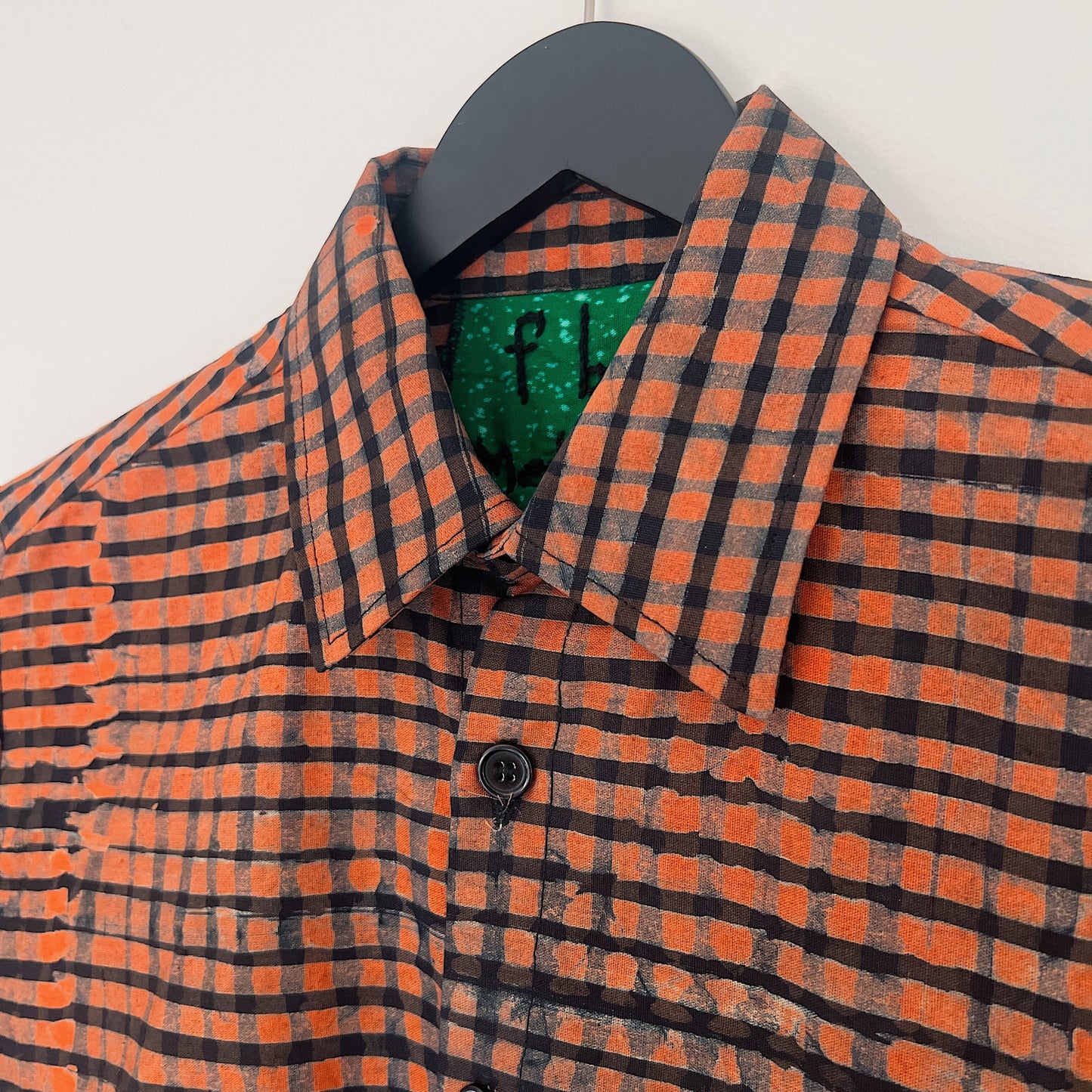 Close-up of the collar and upper chest of the Sudi Shirt in Persimmon Grid, showing the point collar with green Batiki lining, Fahari label, black button, and bold hand-printed persimmon orange and black grid check pattern with natural dye variation

