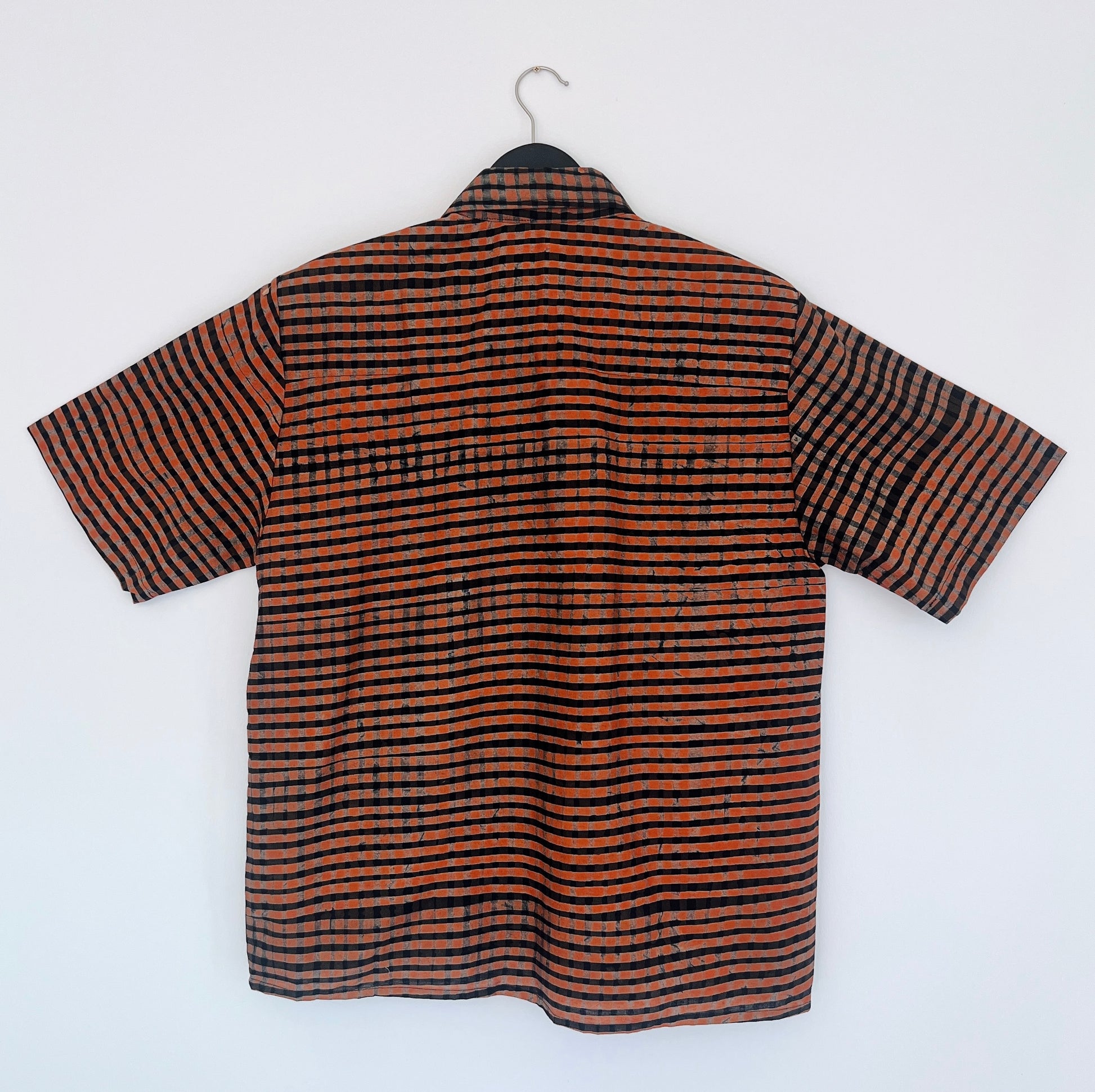 Back view of the Sudi Shirt in Persimmon Grid, hanging on a black hanger against a white wall, showing the relaxed boxy silhouette, short sleeves, and hand-printed persimmon orange and black horizontal grid stripe pattern on 100% cotton Batiki fabric


