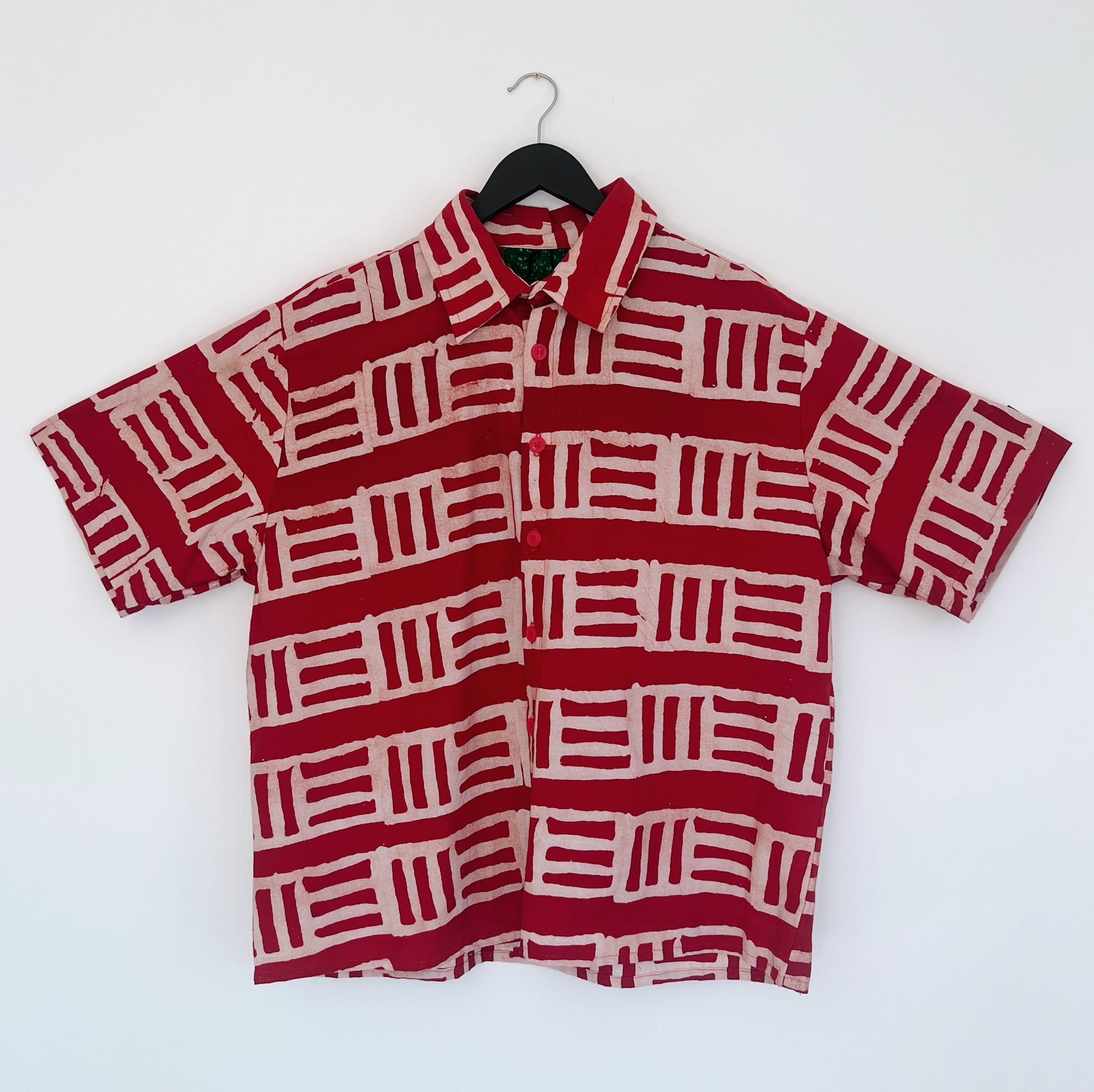 Full front view of the Sudi Shirt in Brick, hanging on a black hanger against a white wall, showing a point collar, short sleeves, button placket, chest pocket, and bold hand-printed brick red and blush geometric block pattern on 100% cotton Batiki fabric

