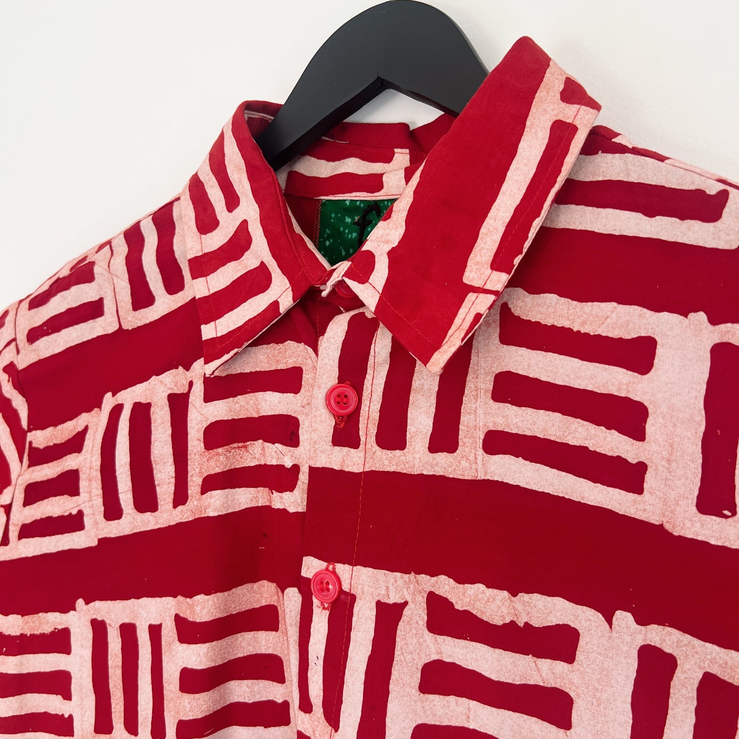 Close-up of the collar and upper chest of the Sudi Shirt in Brick, showing the point collar with green Batiki lining, red button, and bold hand-printed brick red and blush geometric block pattern with natural Batiki dye variation and texture

