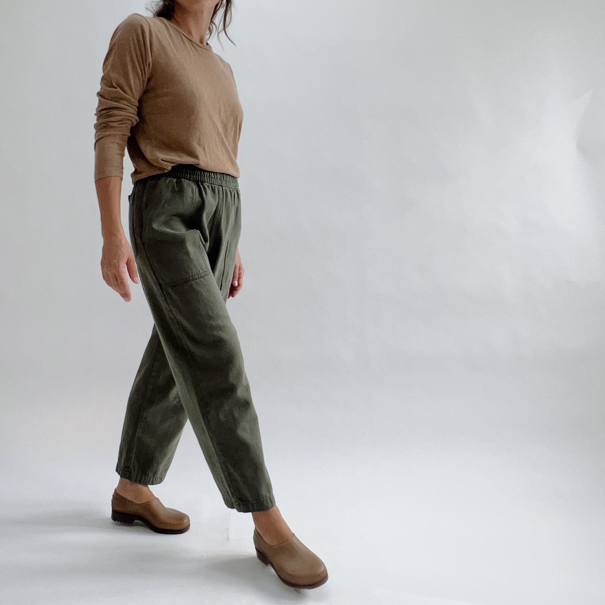 Three-quarter view of model in olive green relaxed pull-on pants with elastic waist and patch pocket, paired with a tan long-sleeve top and brown clogs on a white background.
