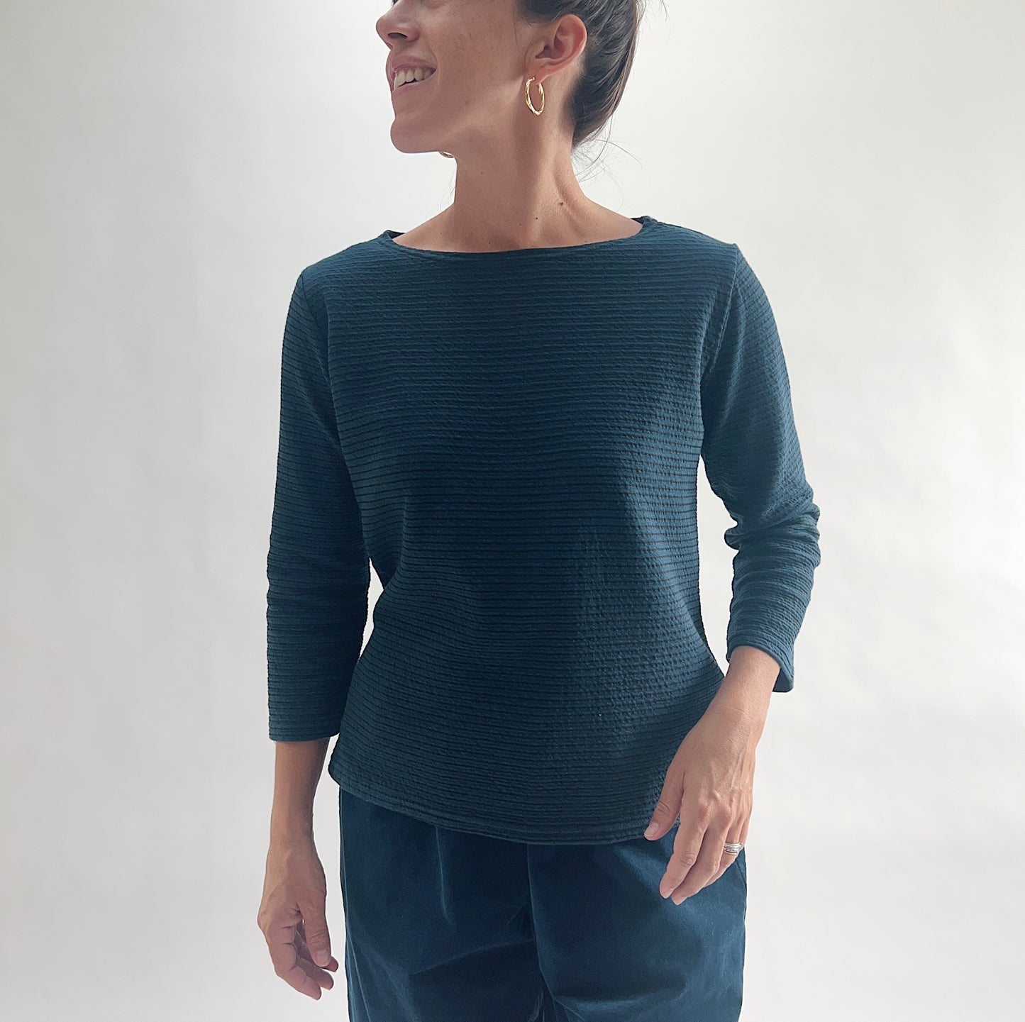 Smiling front view of model wearing a deep teal tonal textured stripe boatneck tee with 3/4 sleeves, showing the wide boatneck neckline and relaxed silhouette, styled with matching teal wide-leg pants.
