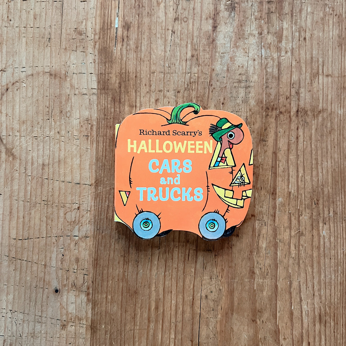 Cover of Richard Scarry's Halloween Cars and Trucks, a die-cut mini board book shaped like an orange pumpkin with blue wheels, featuring Lowly Worm driving a jack-o-lantern car with triangular eyes and a grinning mouth
