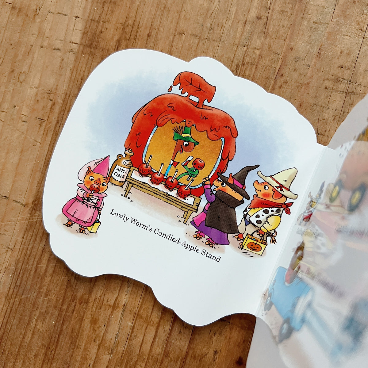 Interior page of Richard Scarry's Halloween Cars and Trucks showing Busytown animal characters in Halloween costumes — a witch, a cowboy, and a fairy — gathered around Lowly Worm's giant candied-apple car, captioned "Lowly Worm's Candied-Apple Stand"
