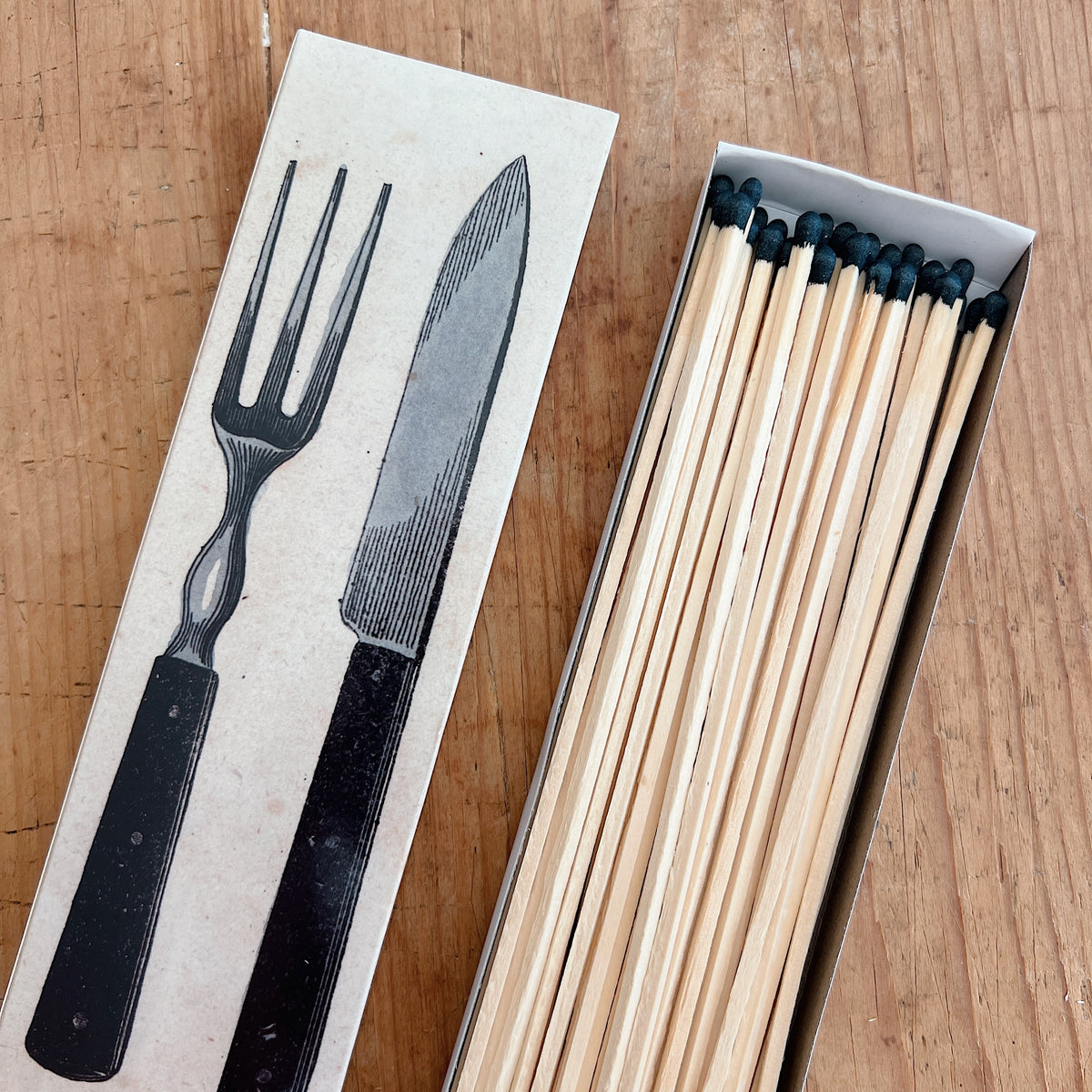 Dotter Silverware Matches box open on warm wood surface, angled top view showing kraft paper box lid with vintage engraving-style black illustration of a large fork and knife, with 50 long wooden matches with black tips neatly arranged in the open box tray
