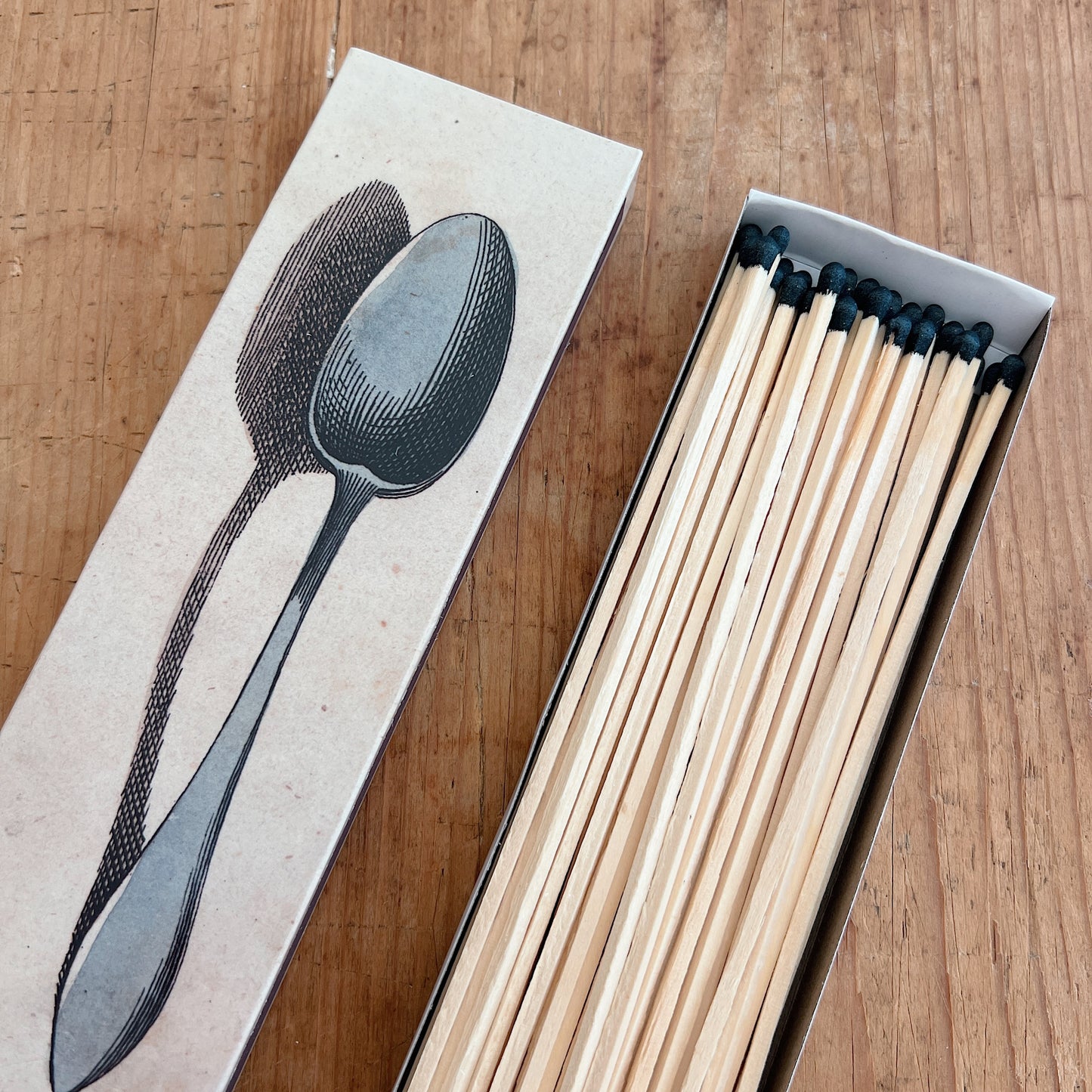 Dotter Silverware Matches box open on warm wood surface, angled top view showing kraft paper box lid with vintage engraving-style black illustration of two spoons, with 50 long wooden matches with black tips arranged in the open box tray
