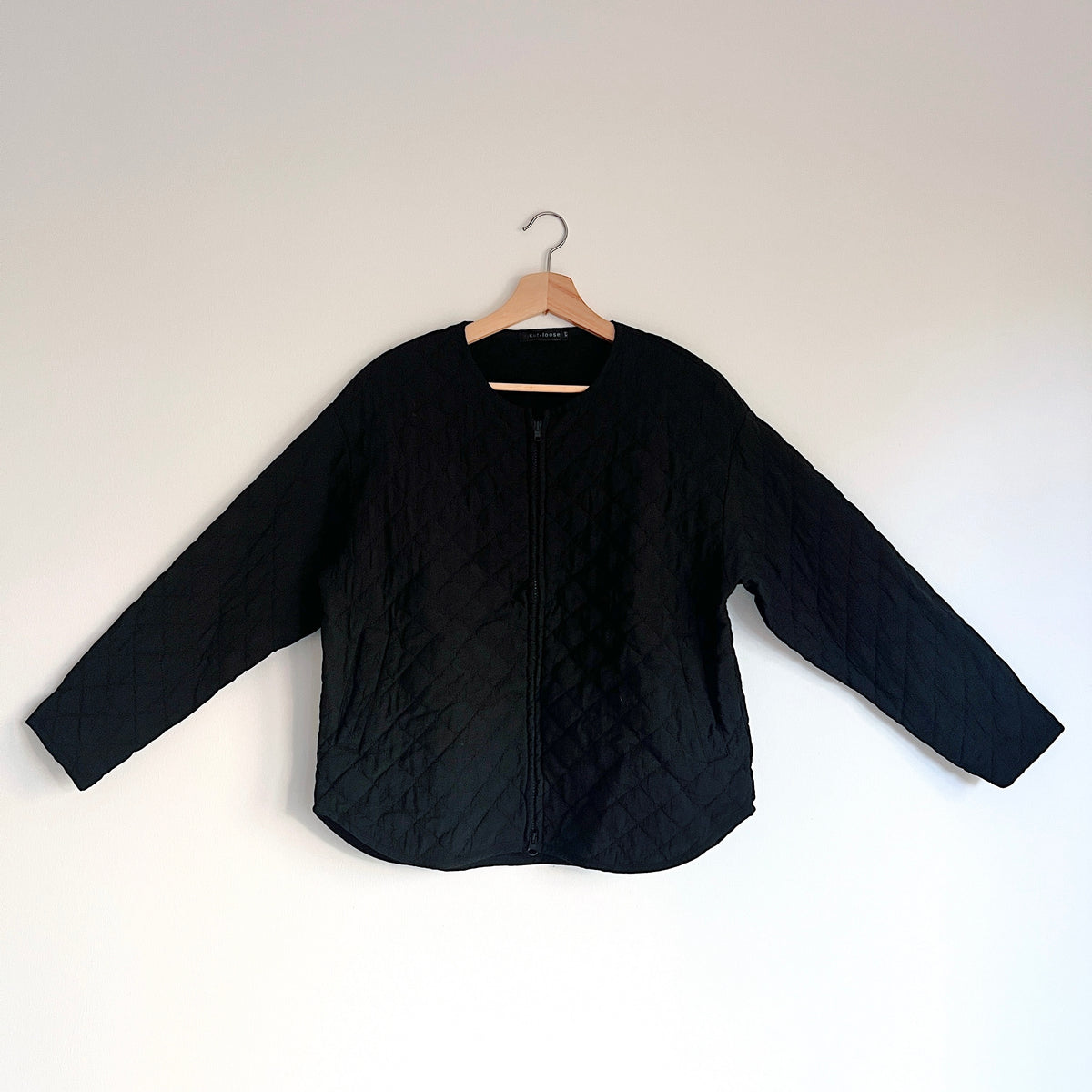 Cut Loose Quilted Bomber Jacket in Black, front view on a hanger showing the zip closure, long sleeves, and curved hem.
