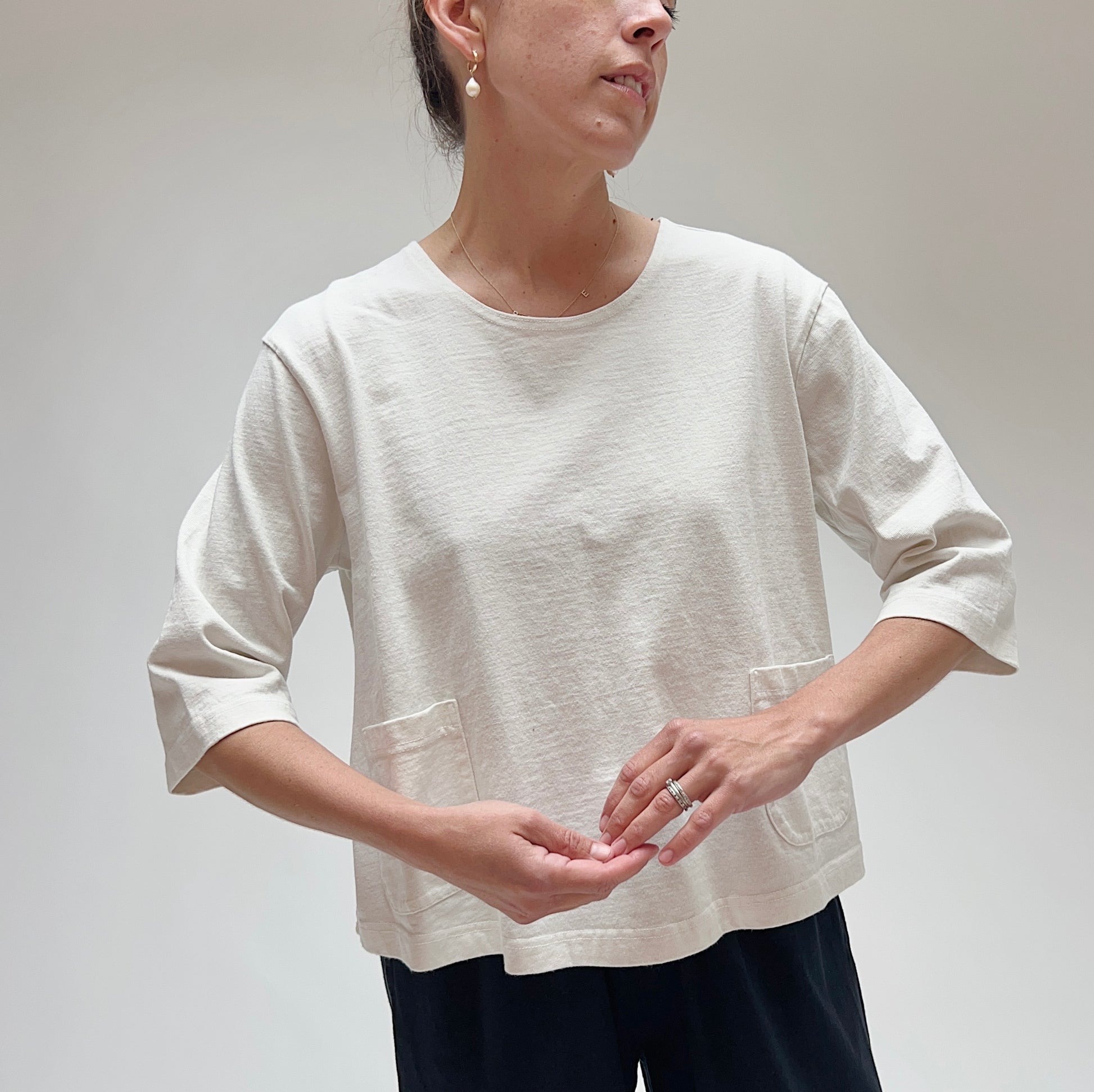 A person wearing a white, 3/4 sleeve cotton top with two front pockets, made by Pacific Cotton.