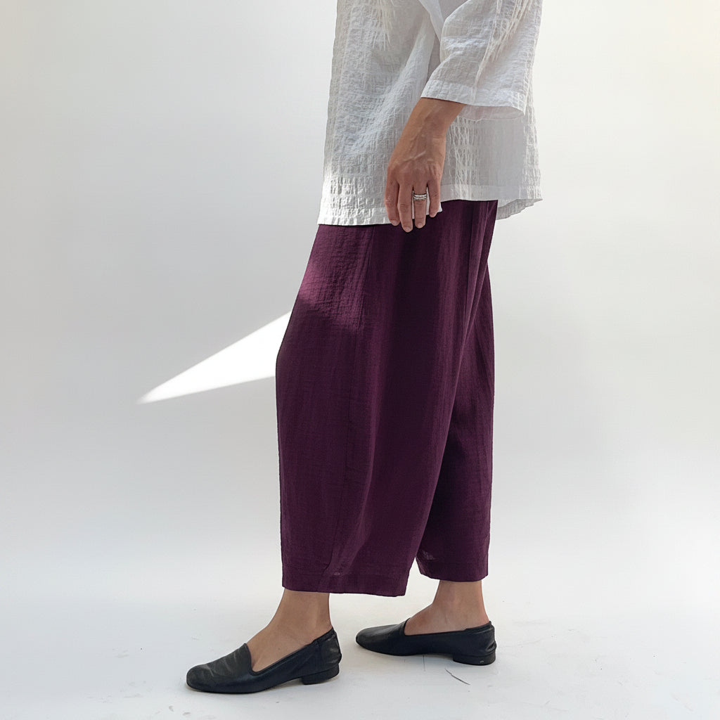 Model wearing Eleven Stitch Crinkle Travel Tucked Hem Pant in Eggplant, side view showing deep eggplant purple crinkle fabric drape, wide-leg balloon silhouette, and tucked gathered ankle hem at cropped length, styled with white textured top and black loafers
