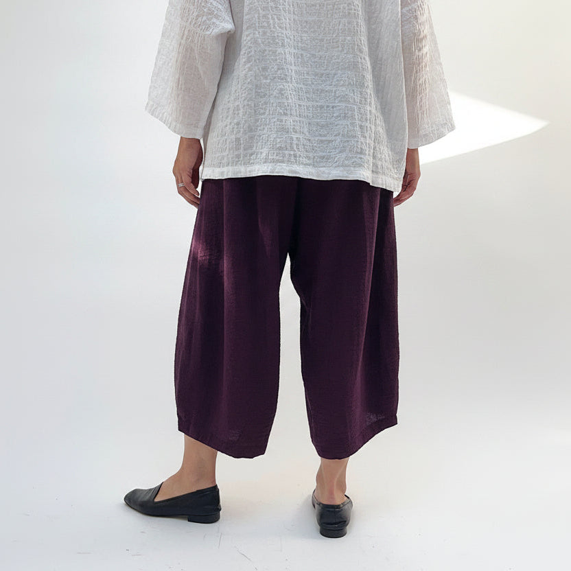 Model wearing Eleven Stitch Crinkle Travel Tucked Hem Pant in Eggplant, back view showing deep eggplant purple crinkle rayon-nylon blend, wide-leg balloon silhouette, elasticized pull-on waistband, and tucked gathered ankle hem at cropped length, styled with white textured top and black loafers
