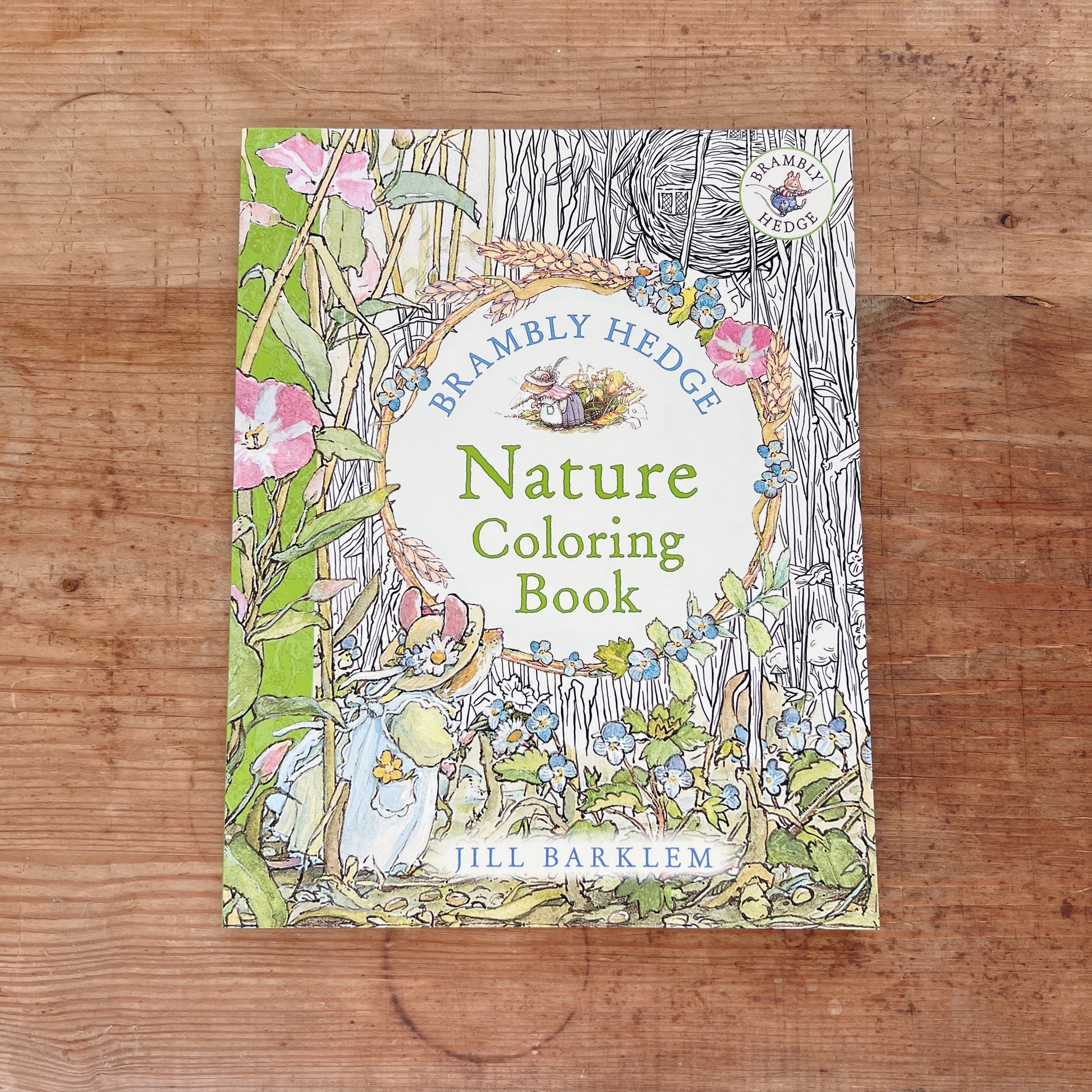 Front cover of the Brambly Hedge Nature Coloring Book by Jill Barklem, featuring a partially colored illustration of a mouse in a blue dress among pink flowers, berry-laden branches, and wheat stalks on the left, transitioning to uncolored black-and-white line art on the right, with the title in green lettering on a white oval and the Brambly Hedge series badge in the upper right corner, displayed flat on a warm wood surface.
