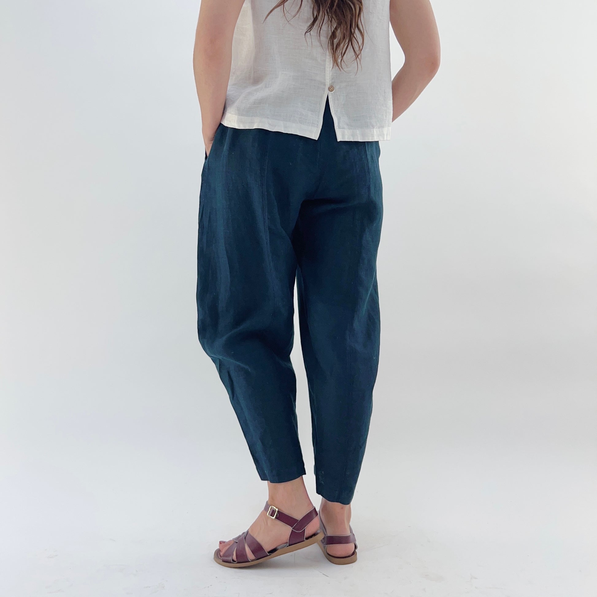 Person wearing navy linen pants and a white linen top on a plain background. They are wearing purple sandals and they are turned away from the camera.