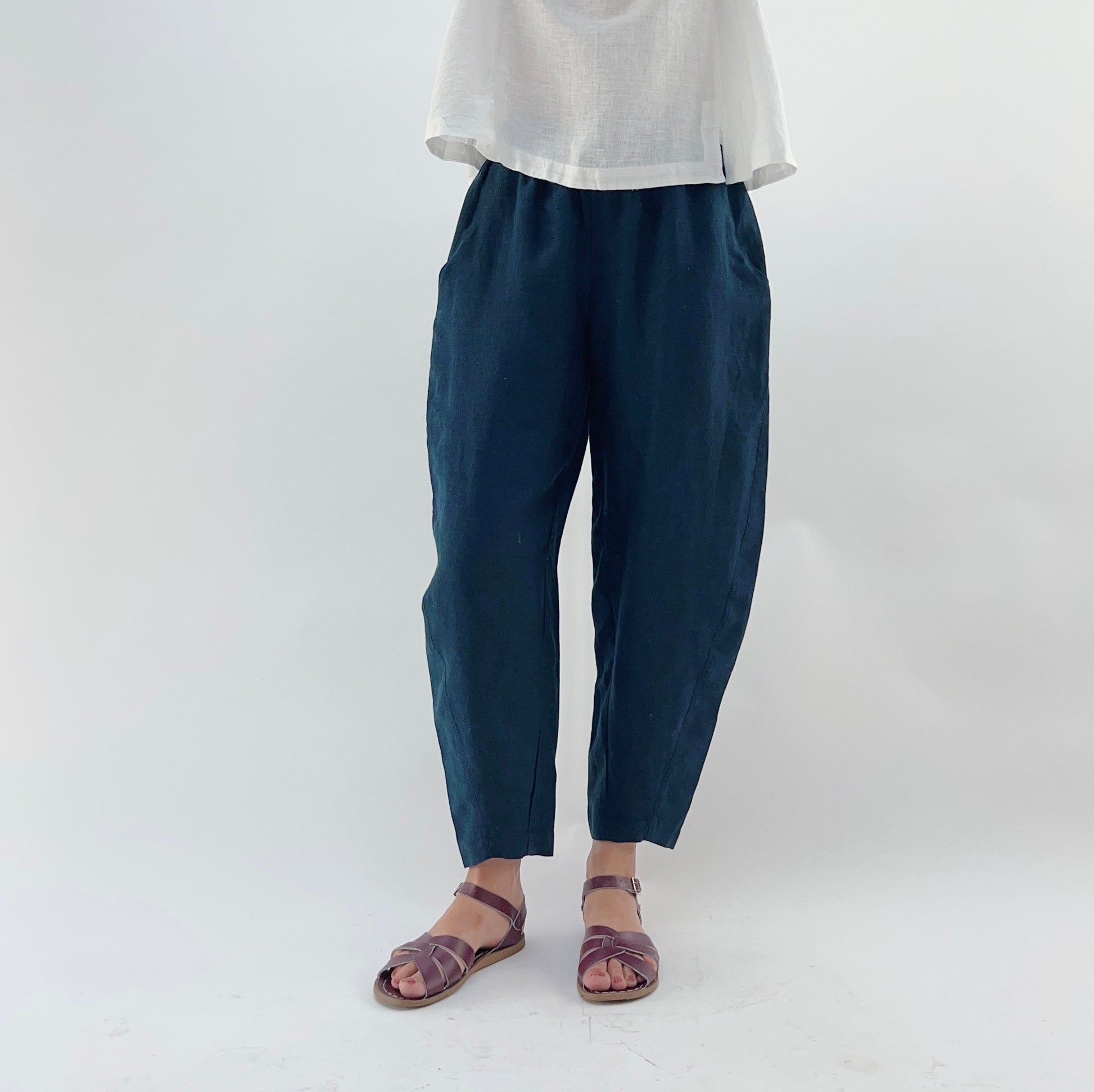 Person wearing navy linen pants and a white linen top on a plain background. They are wearing purple sandals.