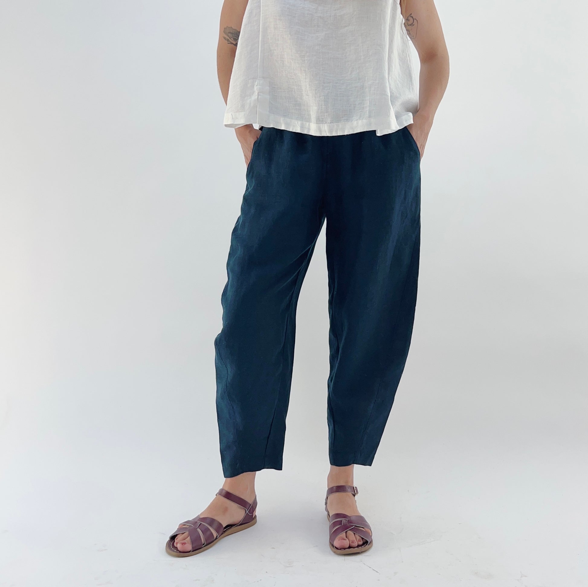 Person wearing navy linen pants and a white linen top on a plain background. Their hands are in their pockets.