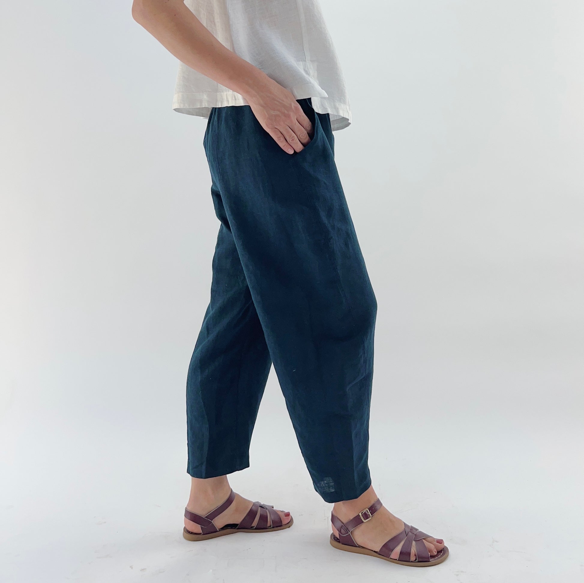 Person wearing navy linen pants and a white linen top on a plain background. They are wearing purple sandals and their hands are in their pockets.