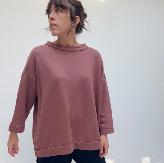 Side-angled front view of a woman wearing the Bryn Walker Alden Fleece Sweatshirt in Tierra, showing the wide draped funnel neck, dropped raglan-style shoulders, wide sleeves with folded cuffs, banded hem, and the relaxed boxy silhouette in dusty rose organic cotton and bamboo fleece, styled with black trousers against a white background
