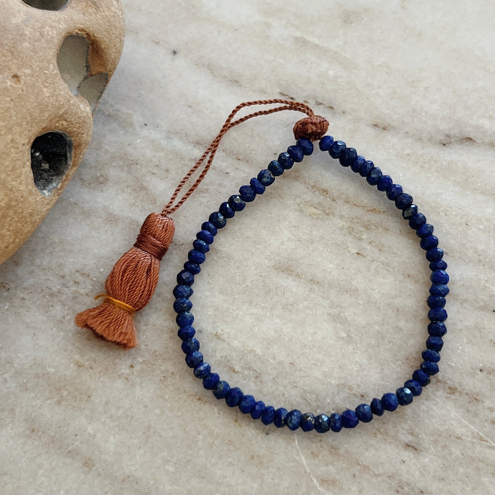 A faceted lapis bead bracelet with an adjustable tassel closure, displayed on a textured surface.