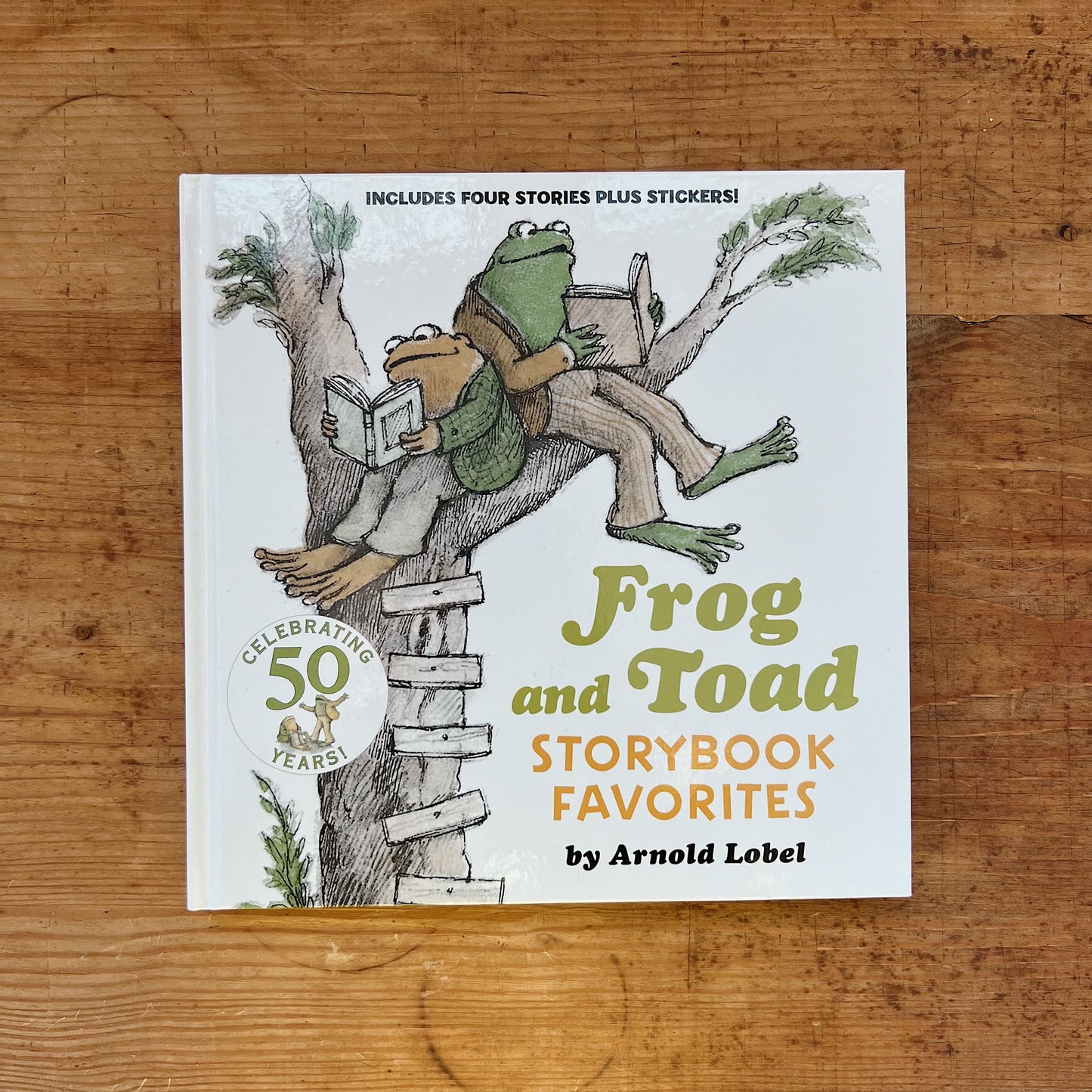 Front cover of Frog and Toad Storybook Favorites by Arnold Lobel, showing Frog and Toad sitting together in a tree reading a book, with a "Celebrating 50 Years" badge and text reading "Includes Four Stories Plus Stickers," displayed flat on a warm wood surface.
