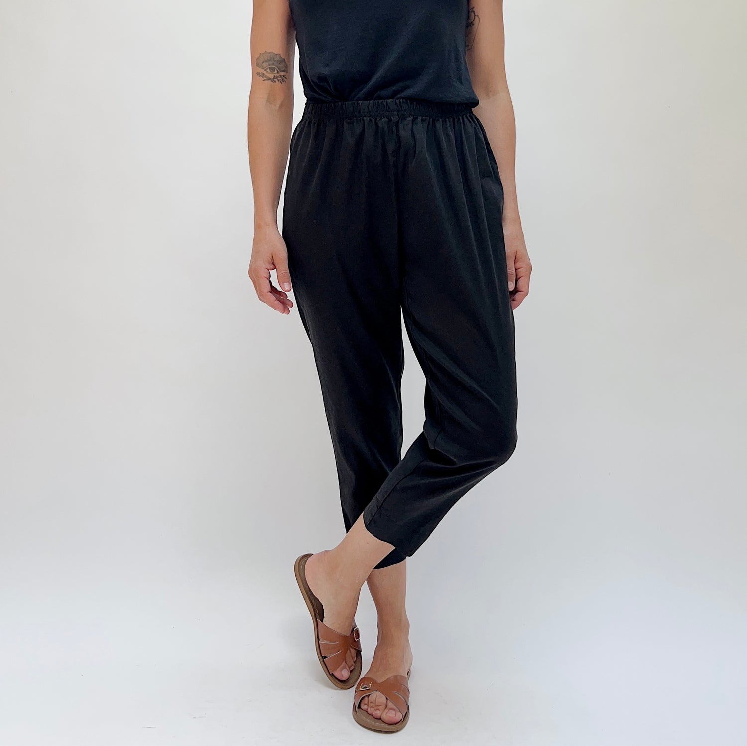 Person wearing black cropped tapered pants and brown sandals with a white background.
