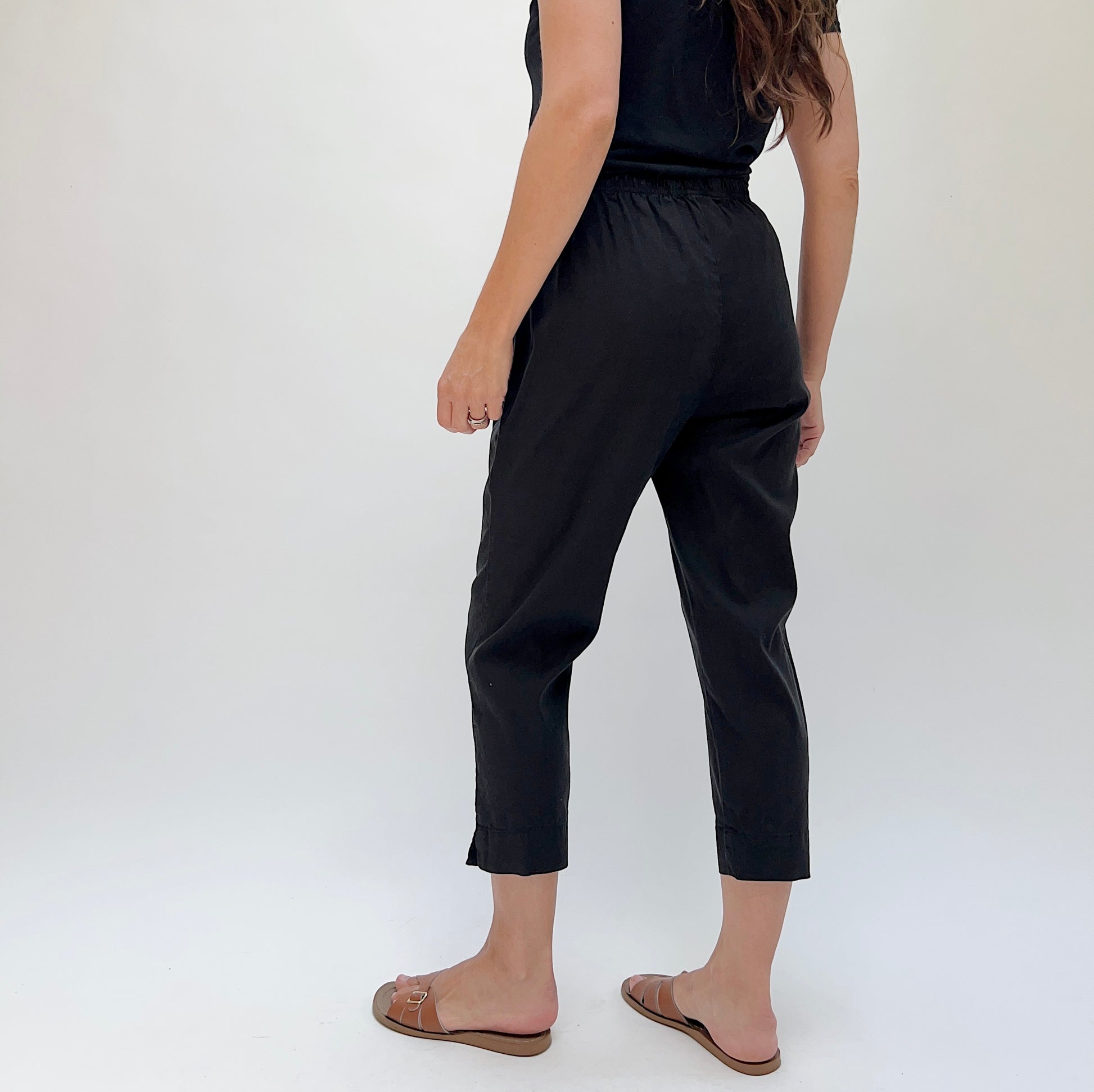 Person wearing black cropped tapered pants and brown sandals with a white background, viewed from behind.