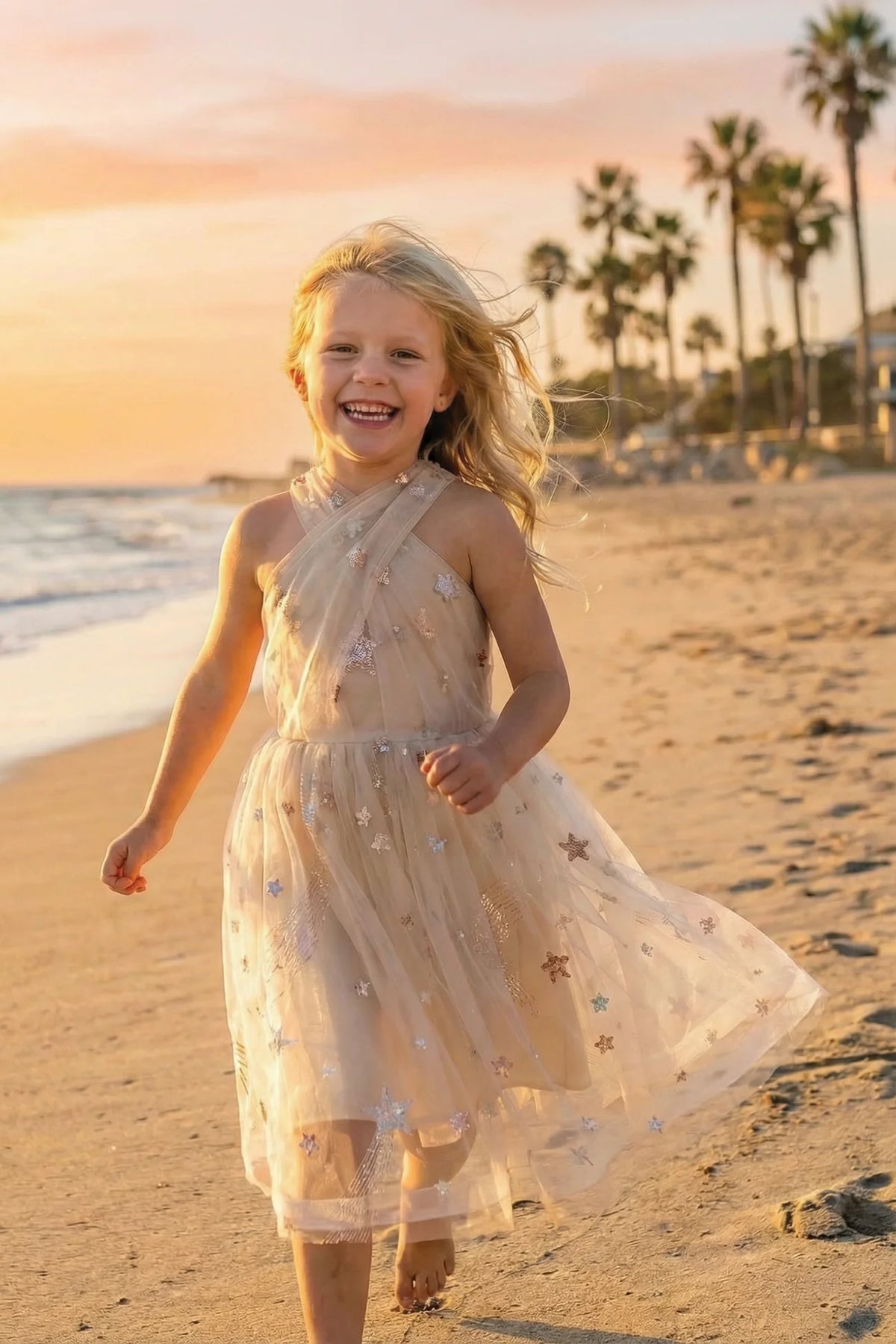 Child wearing the Great Pretenders Eras Dress, a champagne tulle halter dress with crossover neckline and scattered multicolor star sequins, running on a beach at sunset
