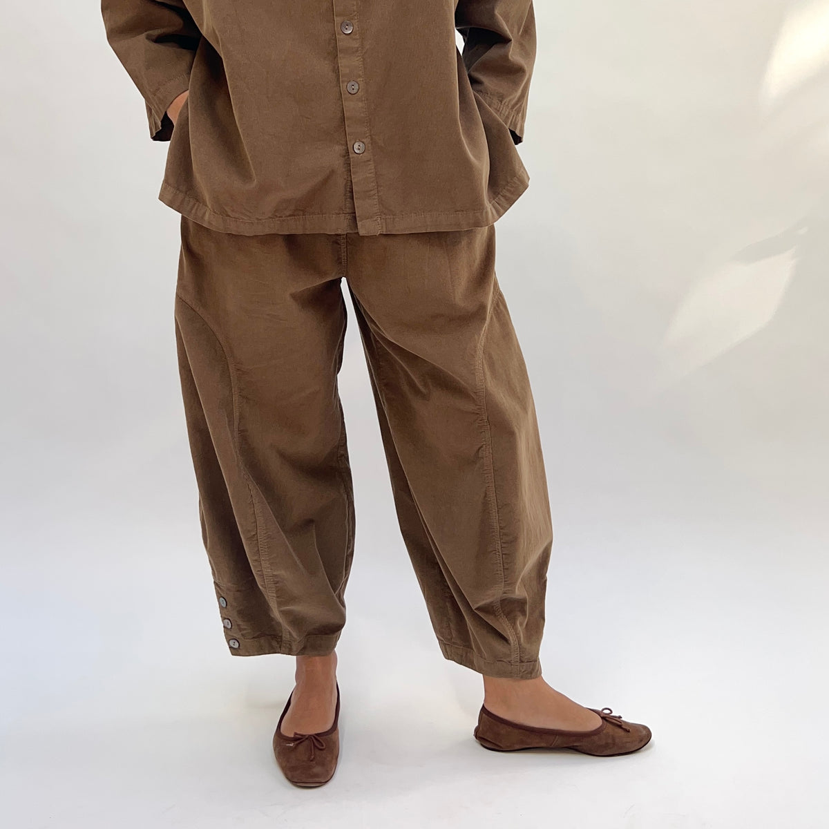 Front lower-body view of a woman wearing the Eleven Stitch Corduroy Doreen Pant in Winter Olive, showing the relaxed high-rise fit, curved balloon leg silhouette, two side pockets, gathered ankle with three shell buttons at the cuff, in fine-wale olive cotton corduroy, styled with the matching corduroy shirt and brown ballet flats against a white background
