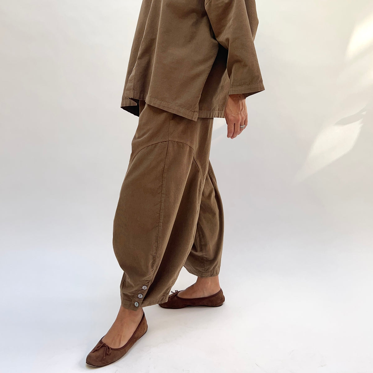 Three-quarter side view of a woman mid-stride in the Eleven Stitch Corduroy Doreen Pant in Winter Olive, showing the curved balloon leg silhouette, curved side seam panel, three shell buttons at the gathered ankle cuff, in fine-wale olive cotton corduroy, styled with the matching corduroy shirt and brown ballet flats against a white background
