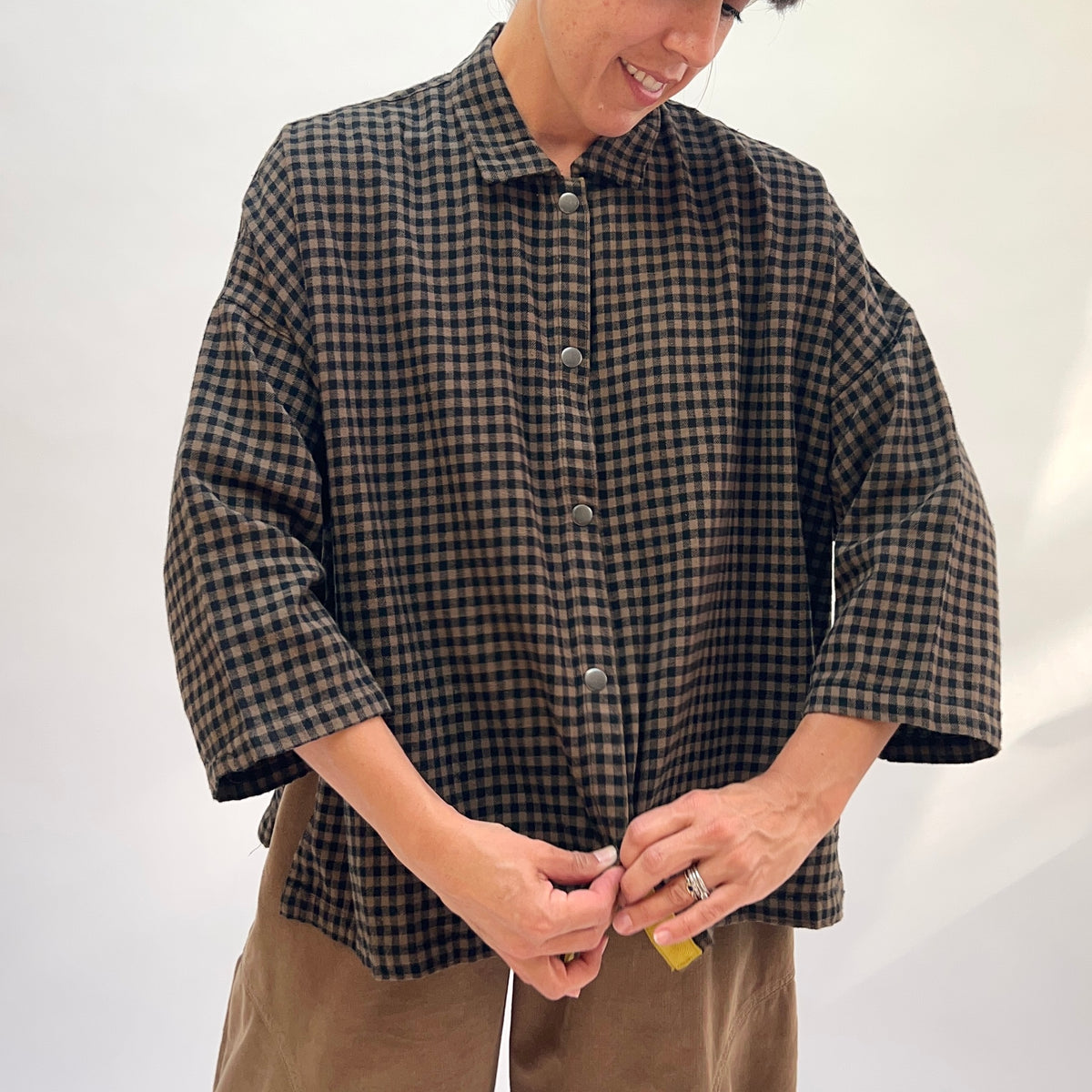 Close front view of a woman wearing the Eleven Stitch Small Check Big Shirt in Winter Olive, showing the point collar, snap button front placket with contrast yellow topstitching, chest patch pocket, and wide 3/4-length sleeves in olive and black small gingham check cotton, styled with tan wide-leg trousers against a white background

