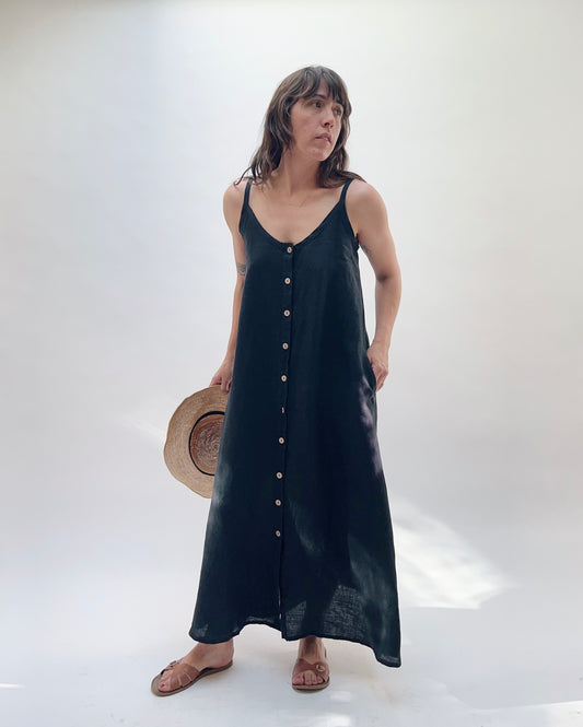 Woman with brown hair wearing a long black linen dress with buttons down the front, holding a tan straw hat on a white background.