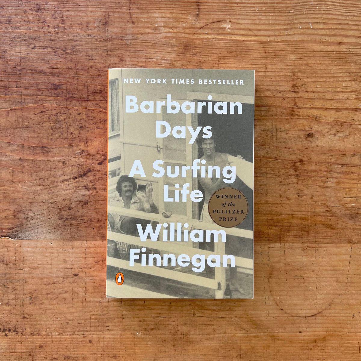 The book 'Barbarian Days: A Surfing Life' by William Finnegan, published by Penguin, is displayed flat on a rustic wooden surface. The cover features a sepia-toned photograph of two men on a surfboard, with bold white text and a gold 'Winner of the Pulitzer Prize' badge.