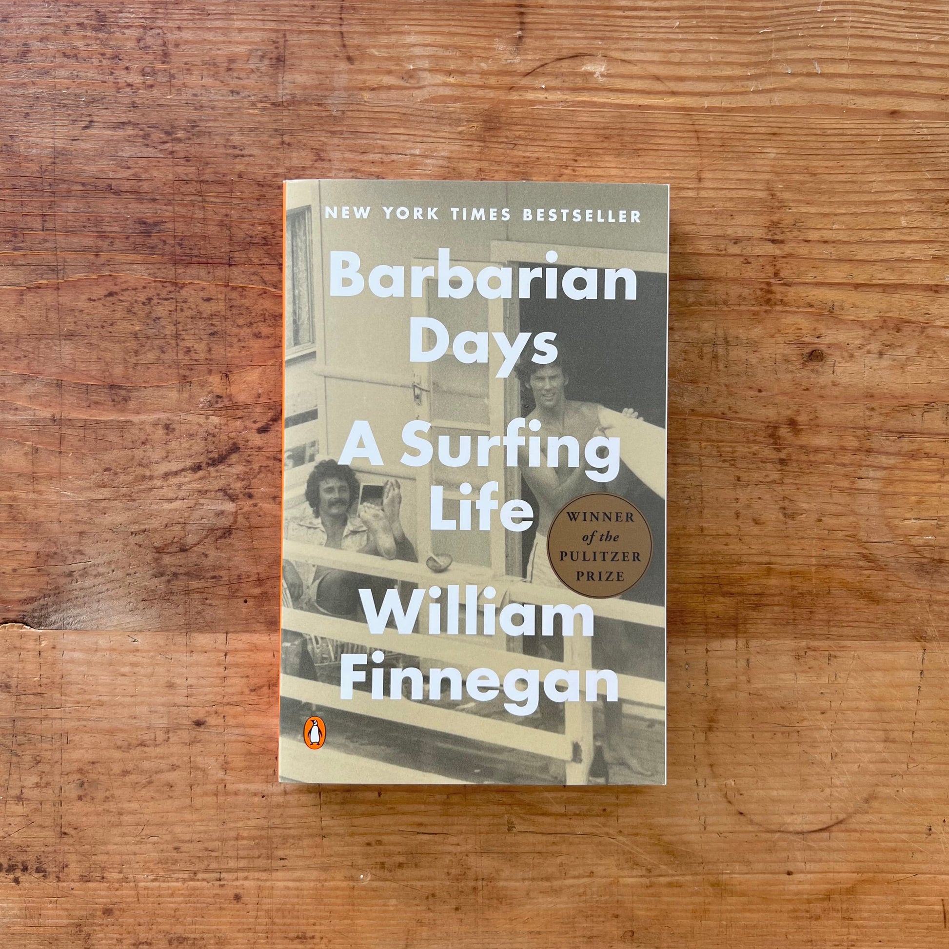 The book 'Barbarian Days: A Surfing Life' by William Finnegan, published by Penguin, is displayed flat on a rustic wooden surface. The cover features a sepia-toned photograph of two men on a surfboard, with bold white text and a gold 'Winner of the Pulitzer Prize' badge.