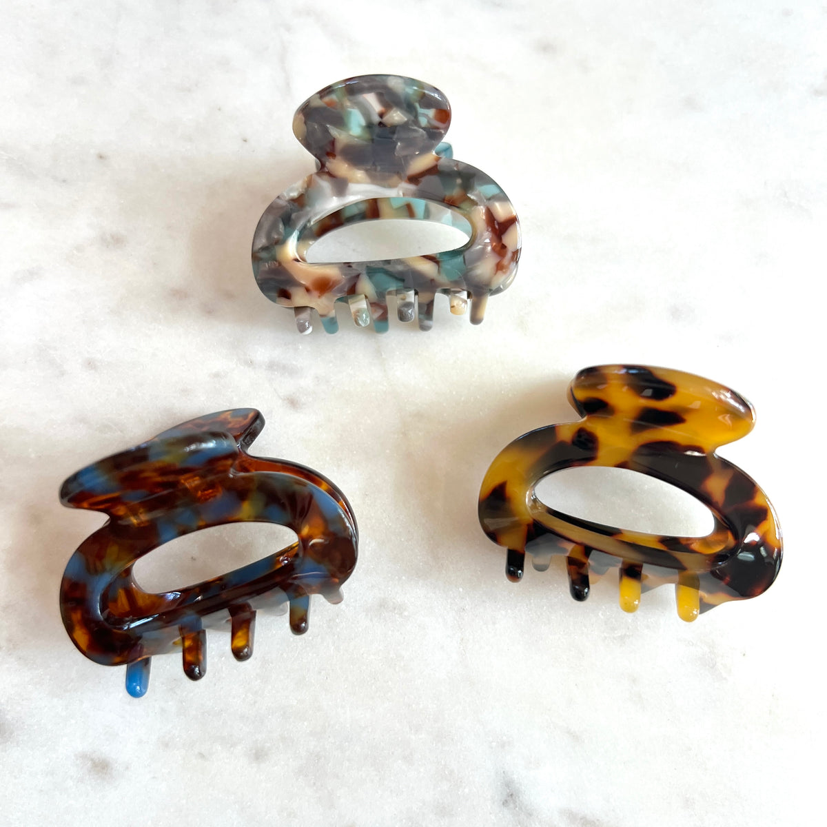 Three tortoiseshell hair claw clips in different patterns and colors, arranged on a white marble surface.