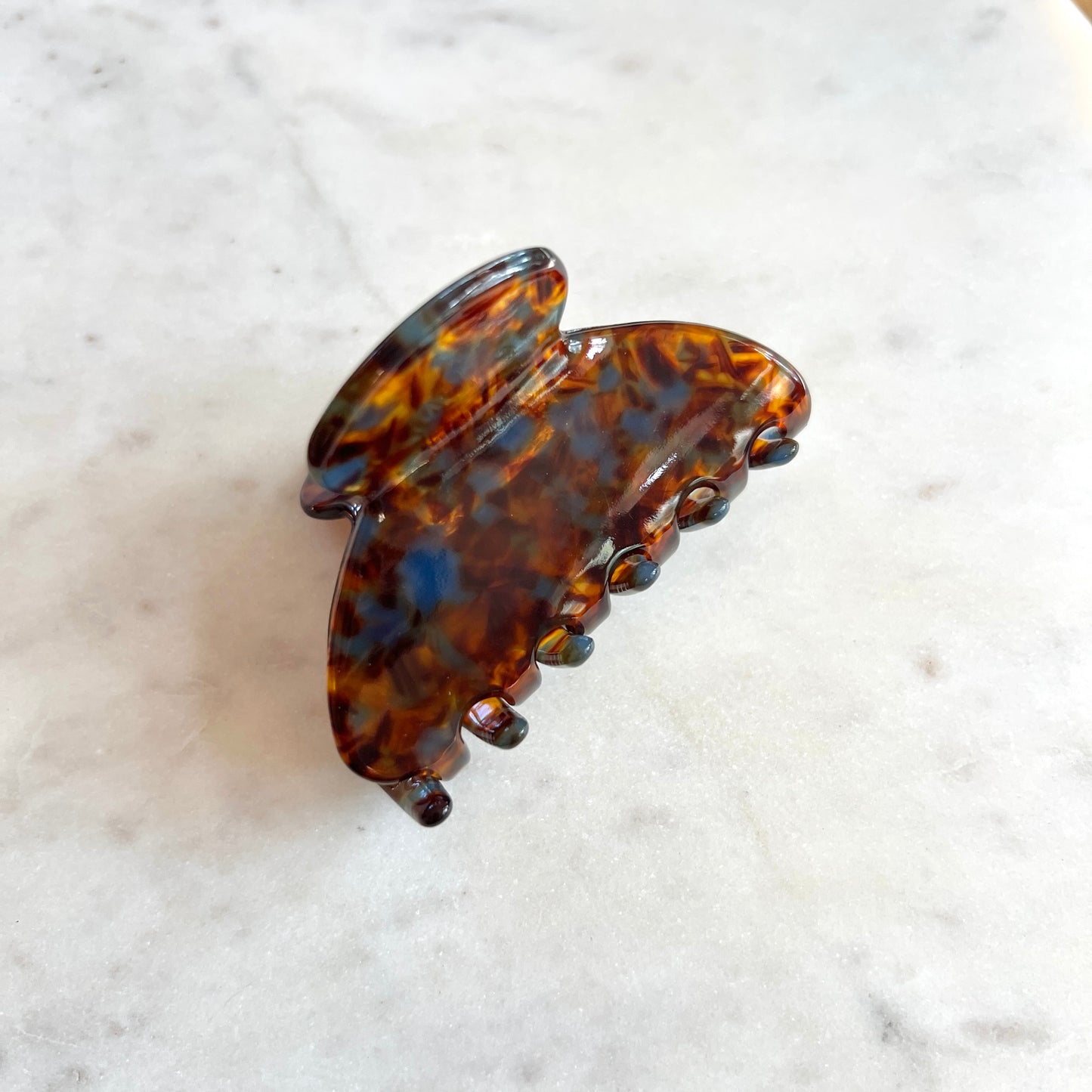 A tortoiseshell hair claw clip with a glossy amber-brown and black tortoise pattern, resting on a white marble surface.