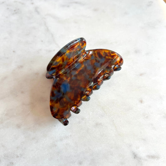 A tortoiseshell hair claw clip with a glossy amber-brown and black tortoise pattern, resting on a white marble surface.