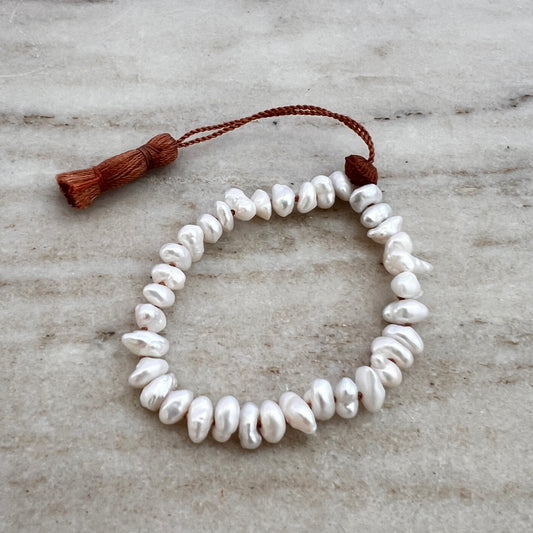 A white pearl tassel bracelet with a brown waxed silk cord and an adjustable tassel closure, displayed on a marble surface.