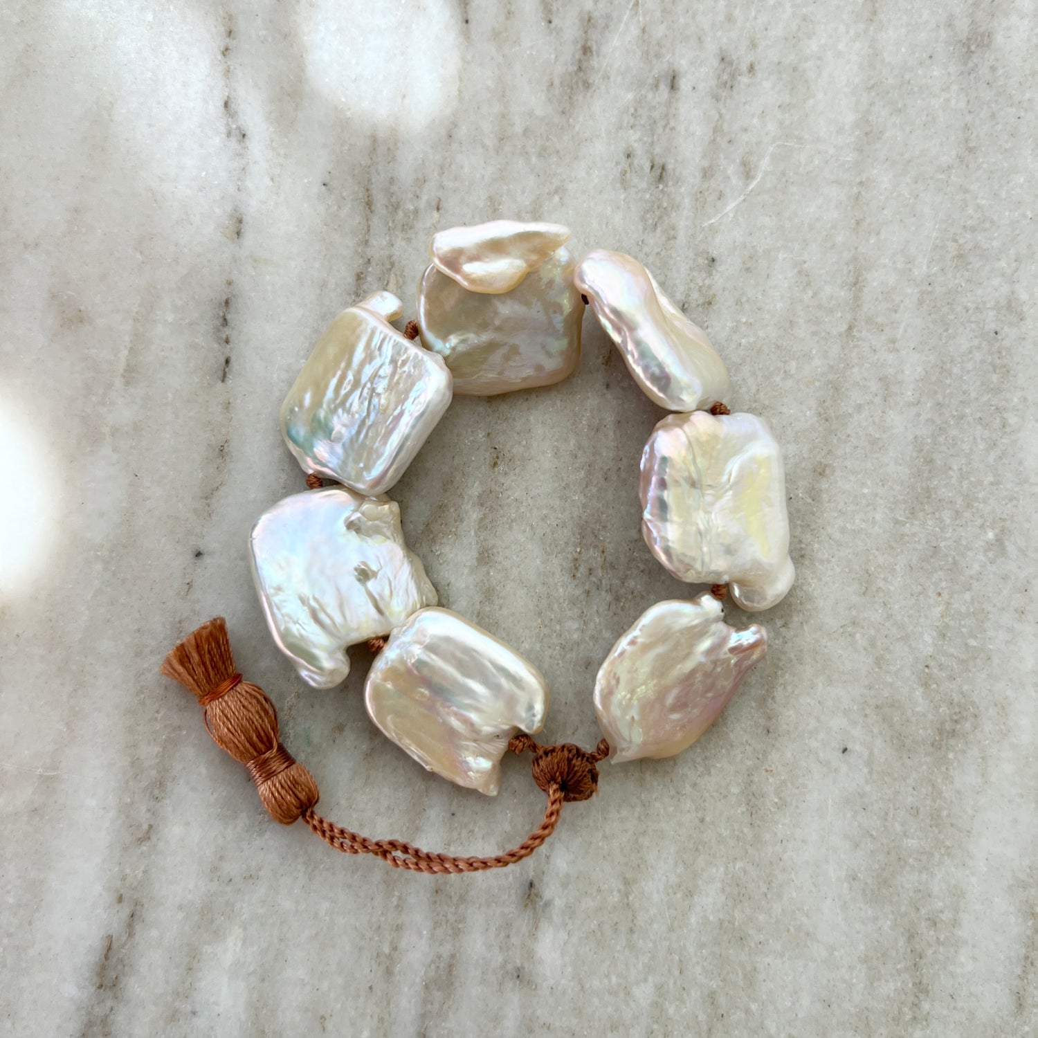 A bracelet with large square pearls on twisted waxed thread with an adjustable clasp and a small tassel on one end, displayed on a marble surface.