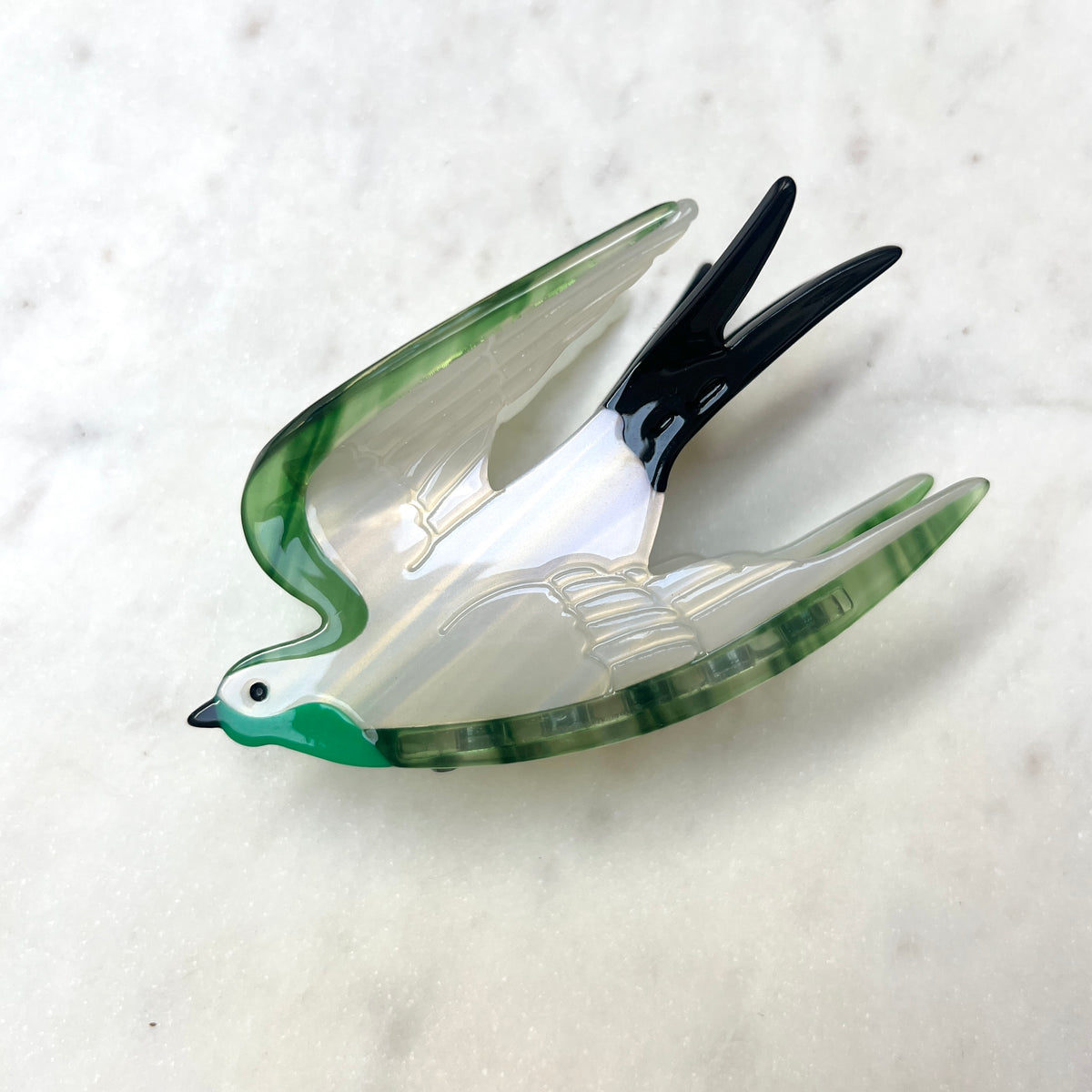 A swallow-shaped hair claw with a glossy white body, green marbled wings and head, and black-tipped wings and tail, displayed on a light marble surface.