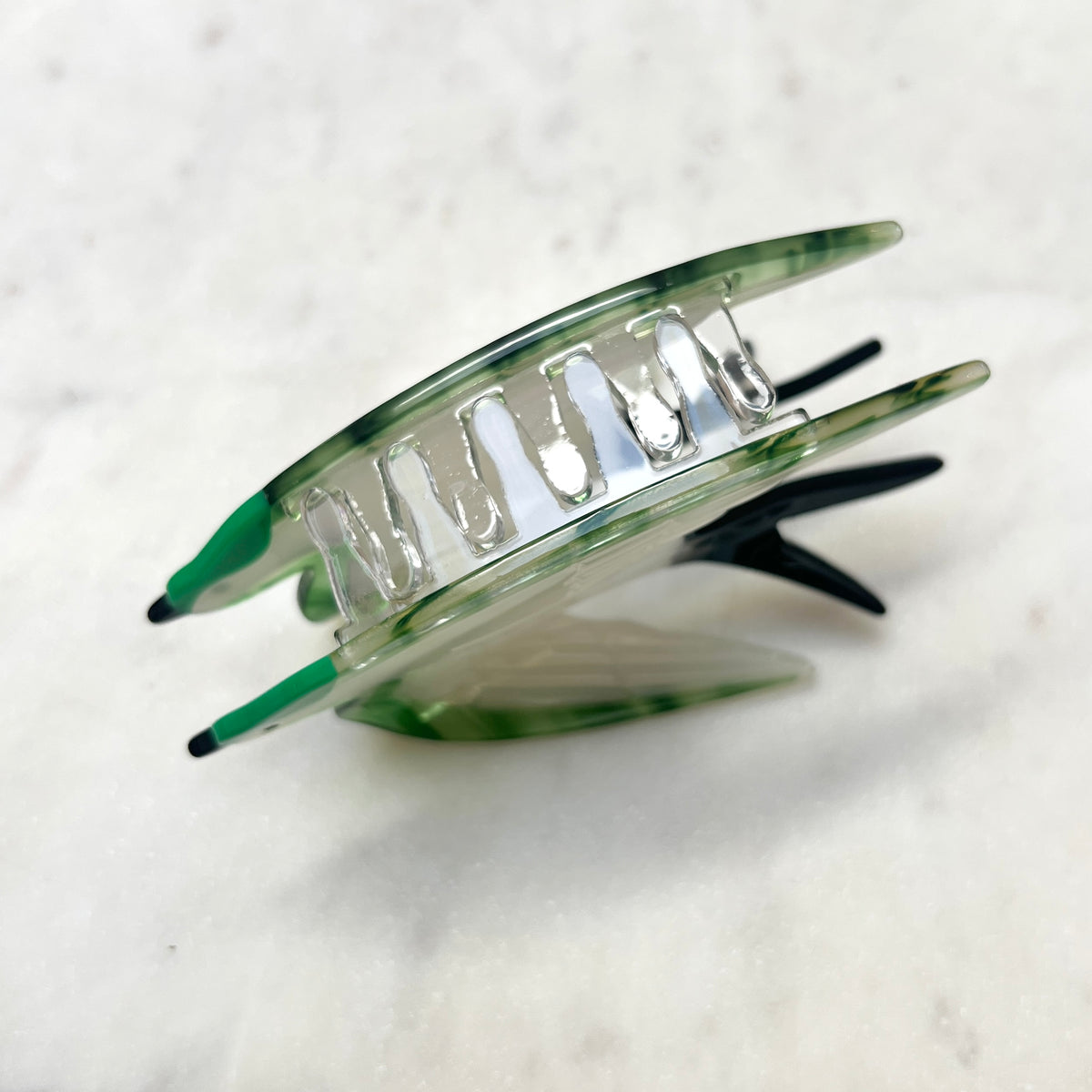 A green and clear acrylic claw hair clip with black metal springs, shown on a light marble surface.