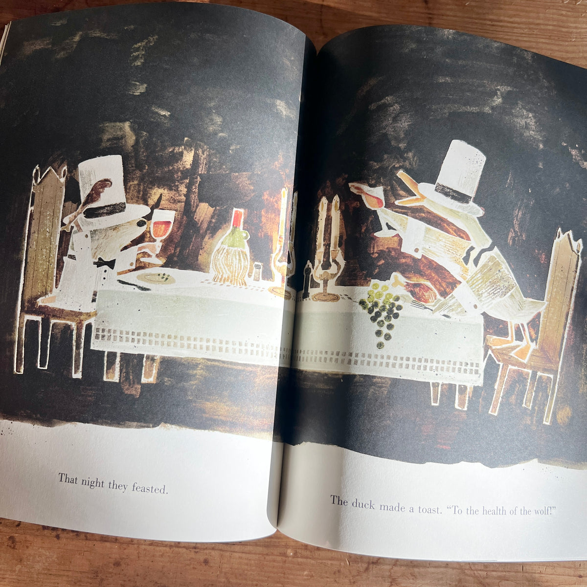 Open pages of The Wolf, the Duck, and the Mouse showing the duck and mouse in white top hats, feasting at a candlelit table inside the wolf’s belly.
