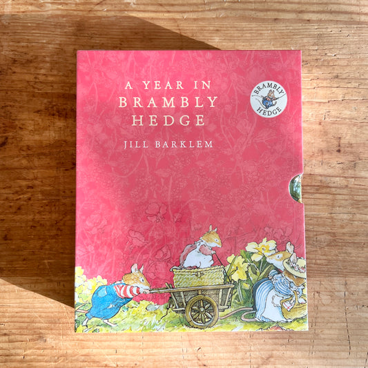 Pink slipcase for A Year in Brambly Hedge by Jill Barklem, illustrated with mice pushing a cart among spring flowers.
