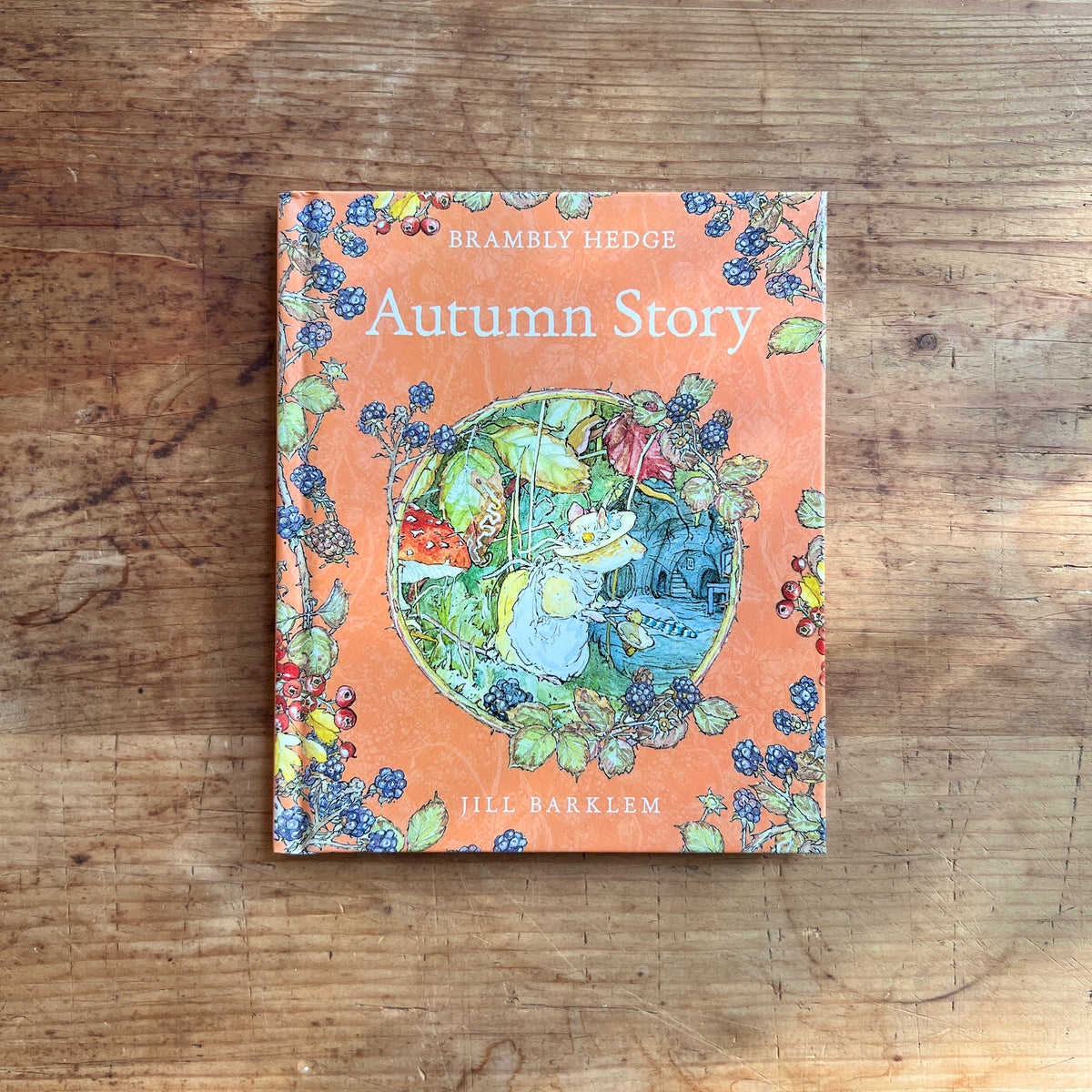 Orange cover of Brambly Hedge Autumn Story by Jill Barklem, showing a mouse beneath blackberry vines, framed by berries and autumn leaves.
