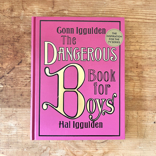 Red hardcover of The Dangerous Book for Boys by Conn and Hal Iggulden, displayed on a wooden table.
