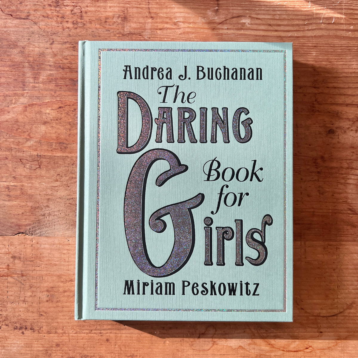 Pale blue-green cover of The Daring Book for Girls by Andrea J. Buchanan and Miriam Peskowitz on a wooden table.
